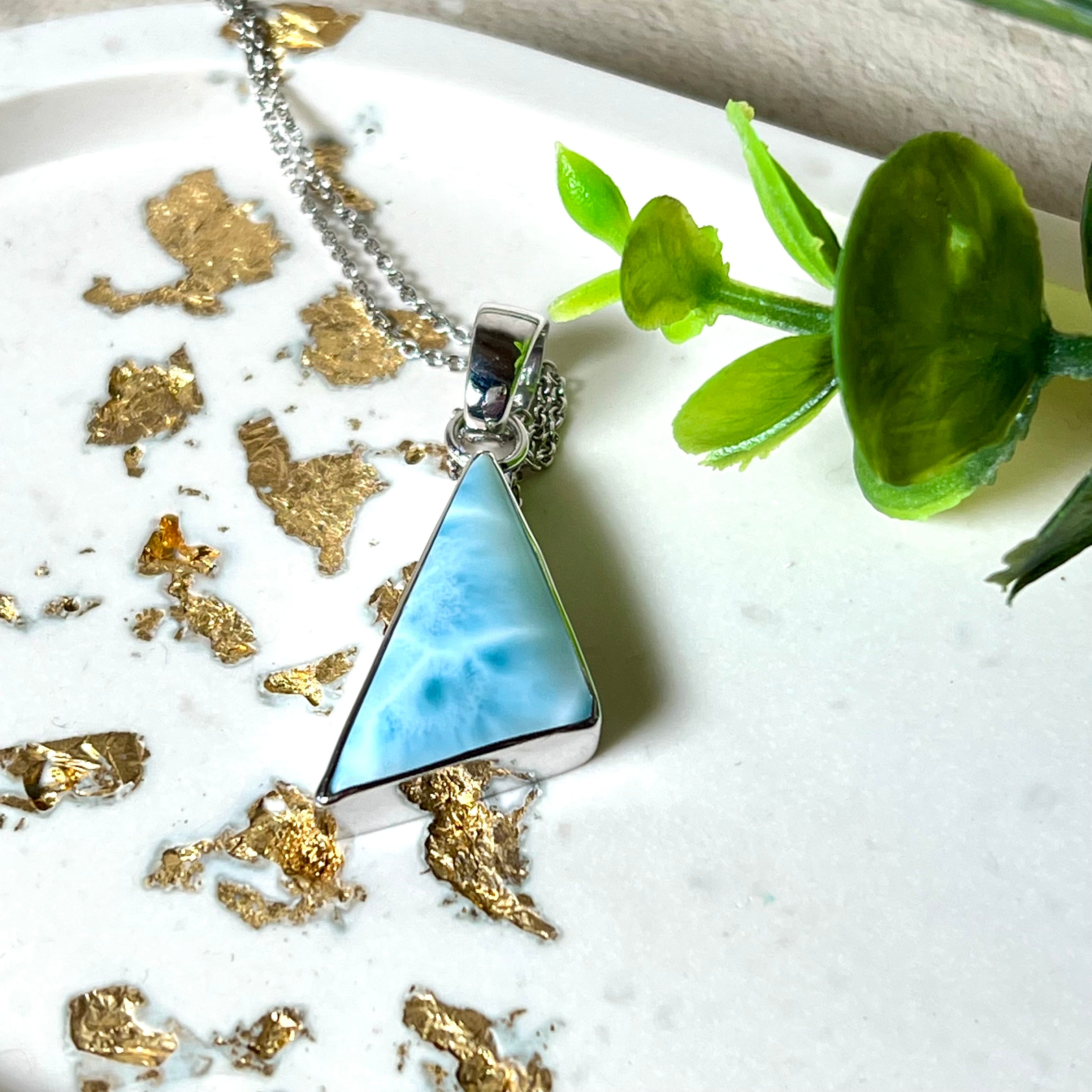 Larimar Dreieck-Anhänger in rhodiniertem 925 Sterling Silber mit filigraner Silberkette, fotografiert auf weißem Untergrund mit Goldflocken und grüner Pflanze – eleganter Edelsteinschmuck mit maritimem Flair.