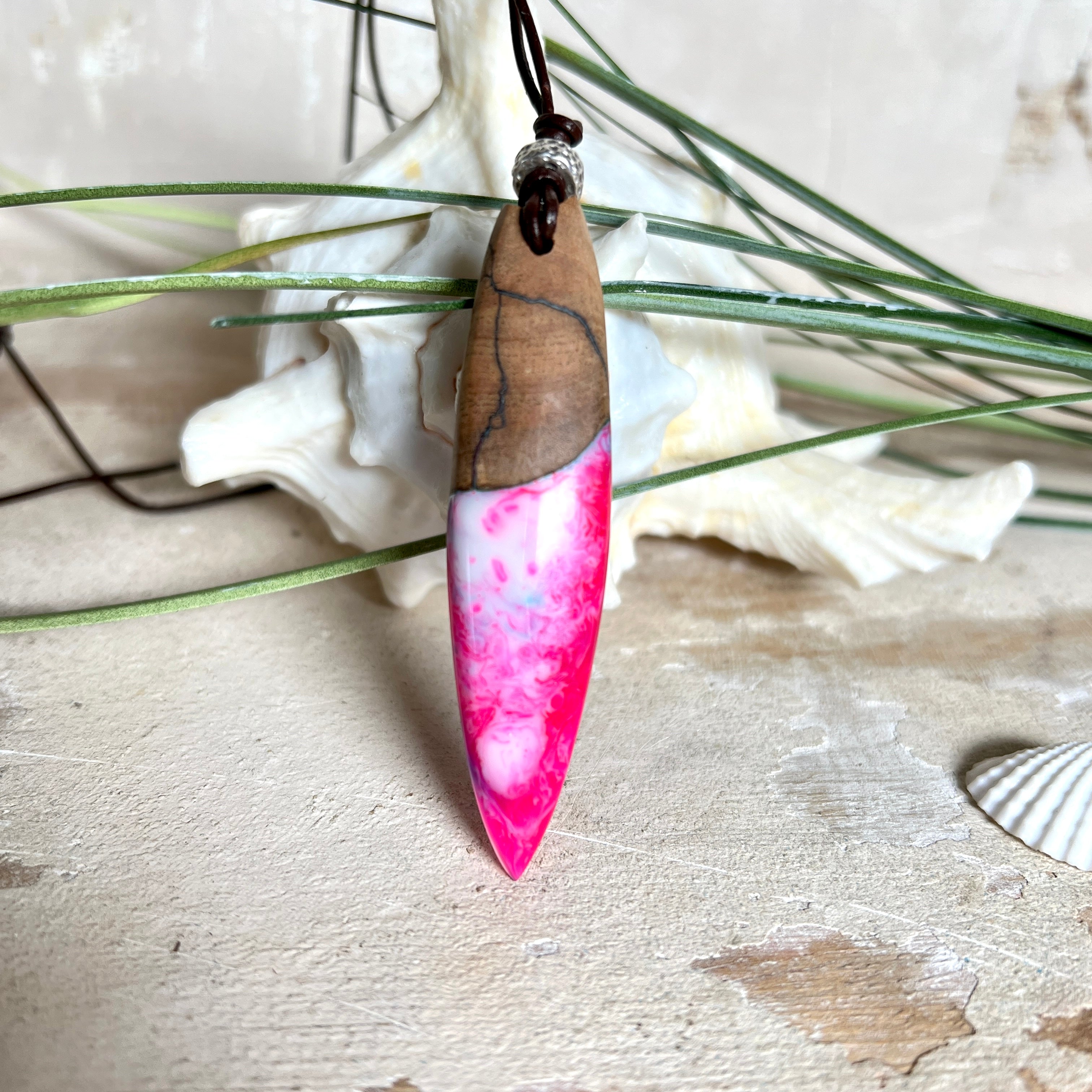 Handgefertigter Surfbrett-Anhänger aus Nussbaumholz und fluoreszierendem Epoxidharz in Pink, Blau und Weiß. Der Anhänger leuchtet zart bei Schwarzlicht und wird an einem verstellbaren Lederband (25–44 cm) mit Holzperlen getragen. Ein maritimes Unikat – ideal für Surfer und Naturliebhaber.