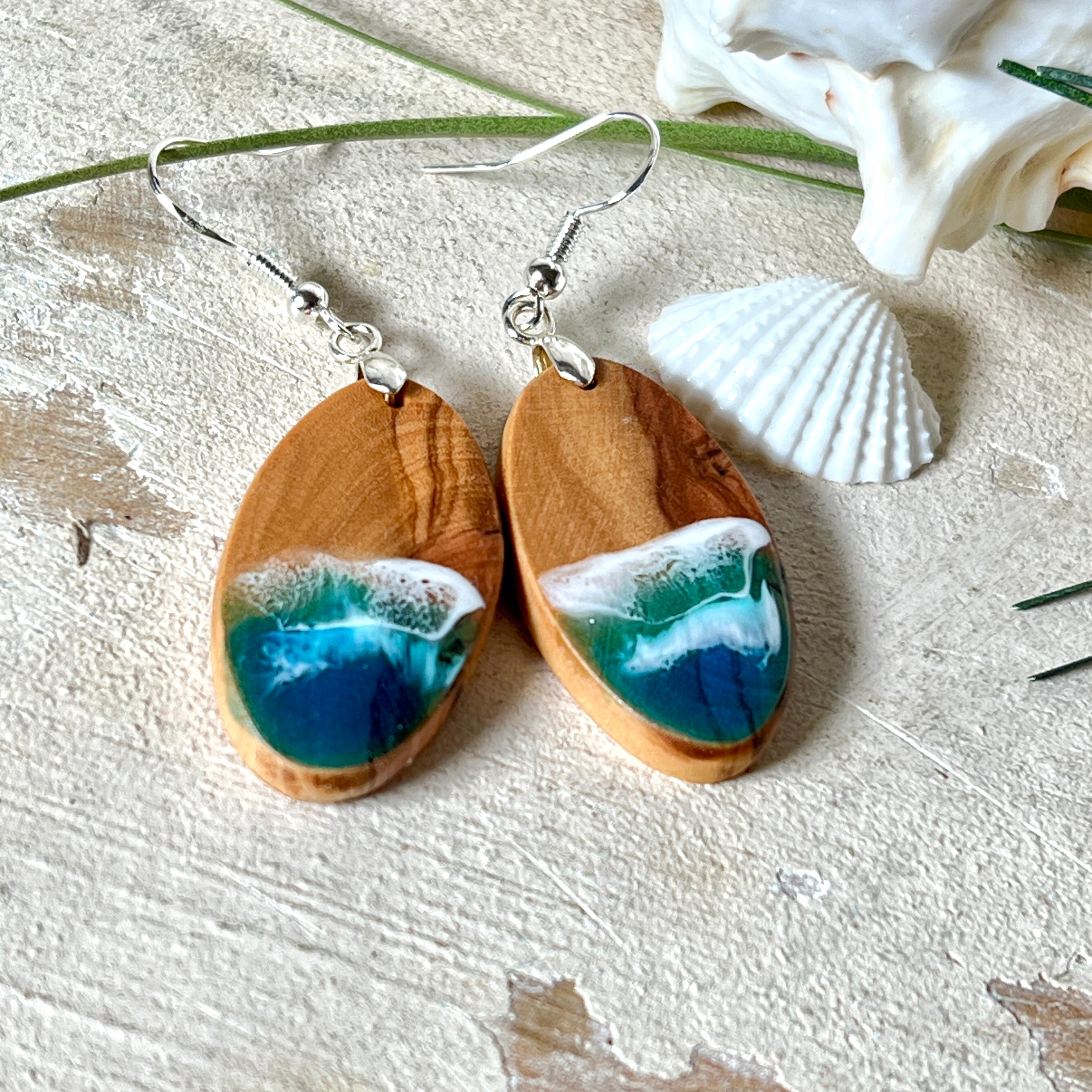 Handgefertigte Ohrringe „Wave Harmony“ aus Olivenholz und Epoxidharz in Blau-, Türkis- und Weißtönen. Das Design erinnert an Meereswellen am Strand. Jeder Ohrring ist ein Unikat mit individueller Maserung. Versilberte, nickelfreie und bleifreie Anhängerschlaufe, 925 Silber Ohrhaken. Maritimer Schmuck – perfekt für Meerliebhaber und als edles Geschenk.