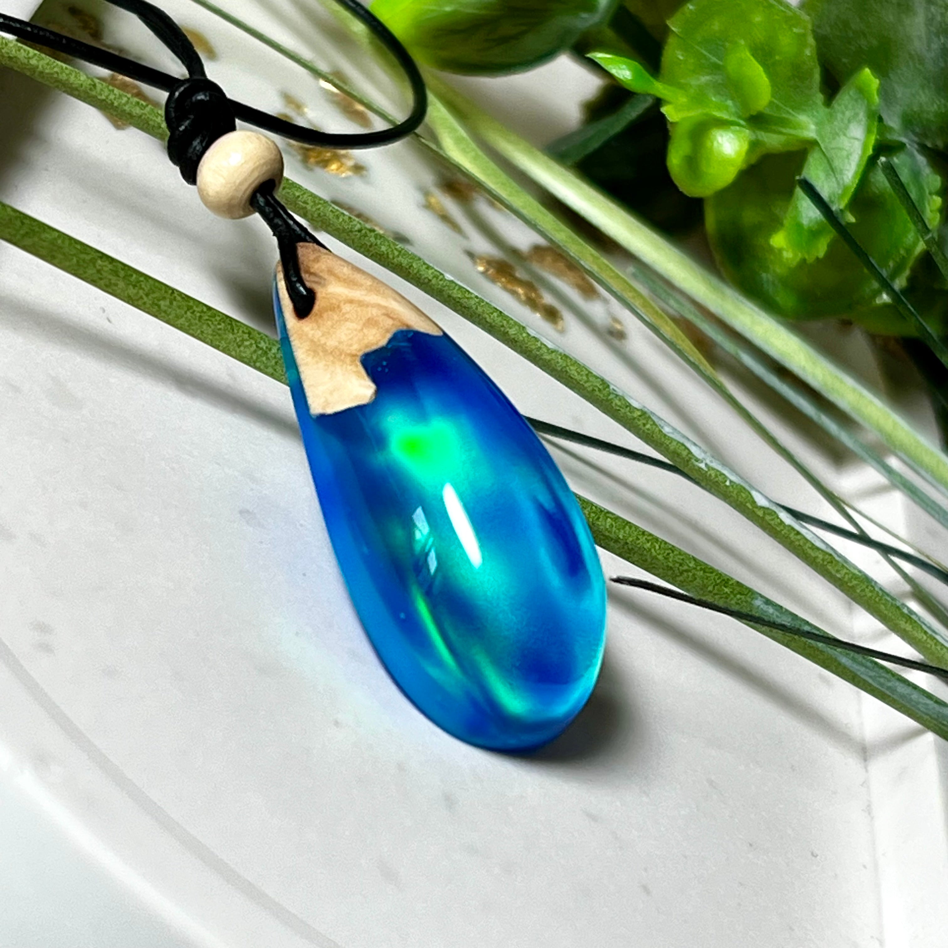 Handgefertigter Tropfenanhänger aus Maserholz und Harz mit synthetischem Opal. Die Farben schimmern je nach Lichteinfall in leuchtendem Blau und Grün – inspiriert von Polarlichtern. Der Anhänger ist ca. 3,9 × 1,8 cm groß und hängt an einem verstellbaren Lederband mit Holzperlen. Jedes Stück ist ein Unikat mit einzigartiger Holzmaserung. Ideal als Geschenk oder stilvoller Begleiter im Alltag.