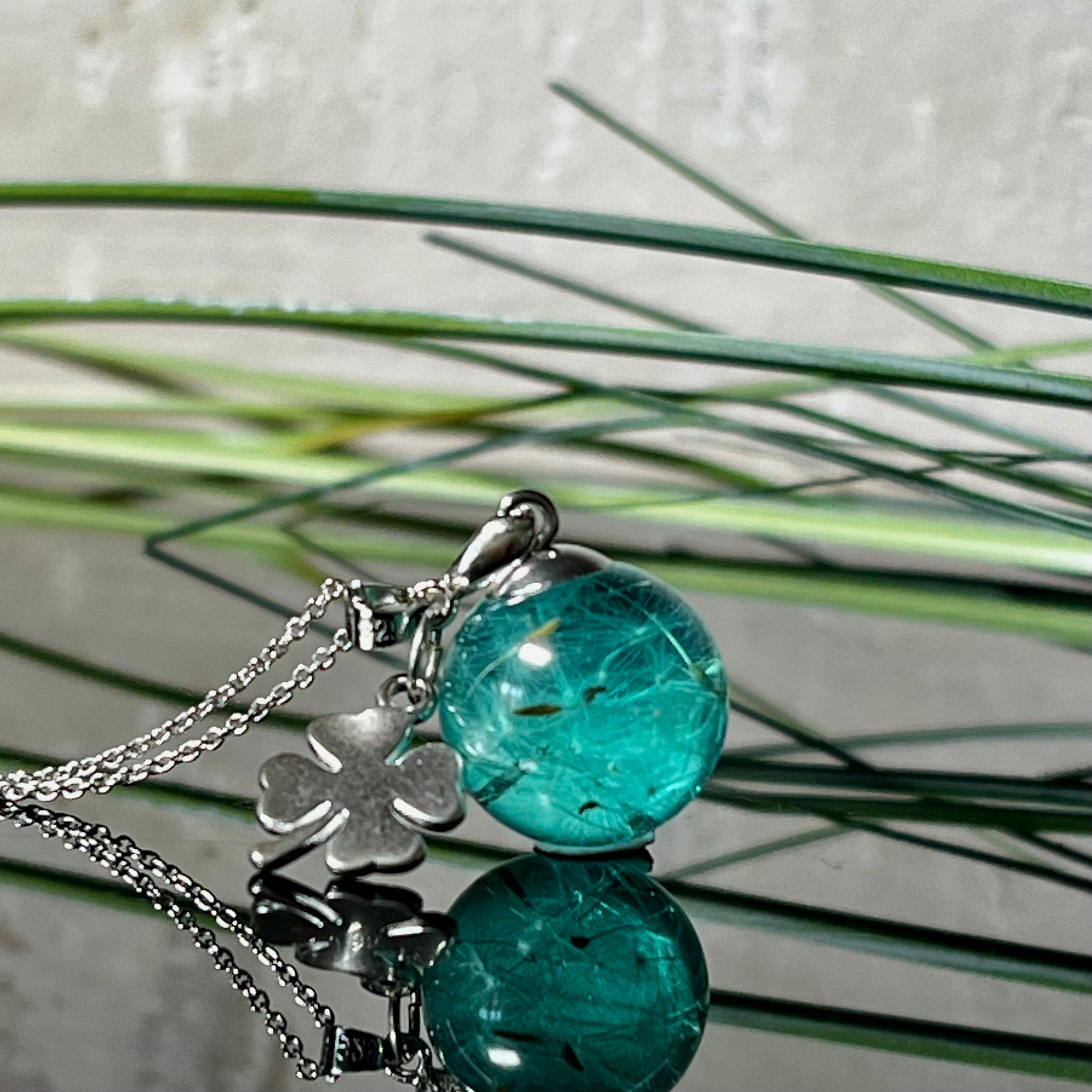 Eine handgefertigten Halskette mit türkiser Harzkugel, in der echte Pusteblumensamen eingeschlossen sind. Ergänzt durch einen glänzenden Edelstahl-Kleeblatt-Charm und einer feinen rhodinierten 925 Silberkette. Der Anhänger symbolisiert Glück, Leichtigkeit und neue Wege – ein emotionales Schmuckstück mit Bedeutung.