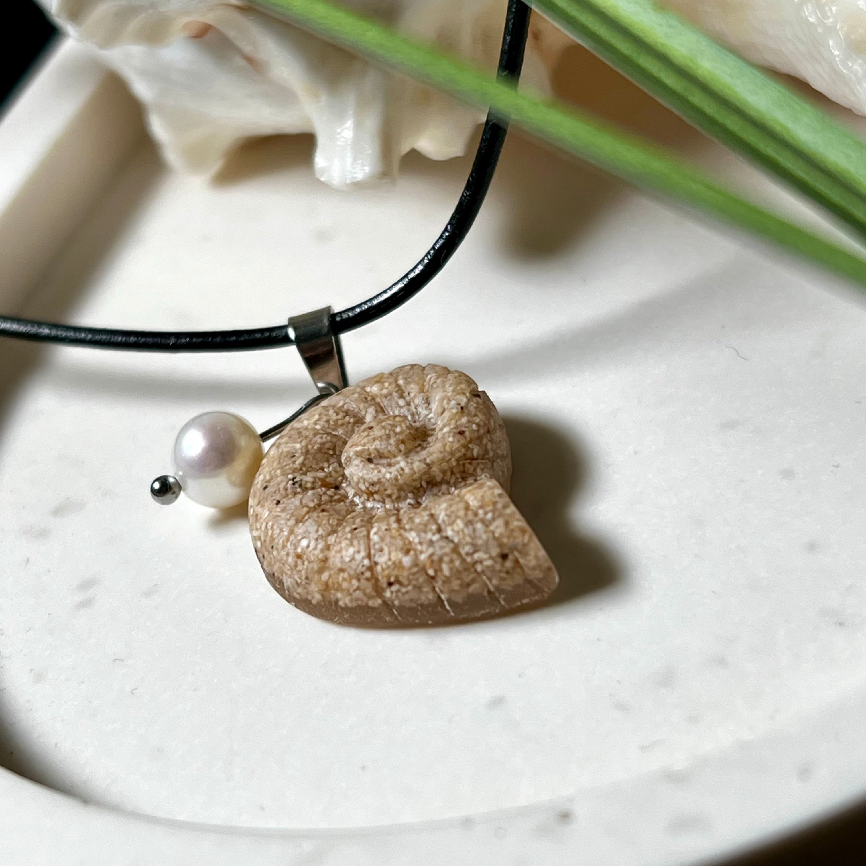 Handgefertigter Ammonit-Anhänger aus echtem Meersand & Harz mit filigraner Edelstahlöse und einer edlen Süßwasserperle. Maritimer Schmuck, inspiriert von der Kraft und Schönheit des Ozeans. Jedes Stück ein Unikat. Präsentiert auf einer Muschel, mit Lederkette.