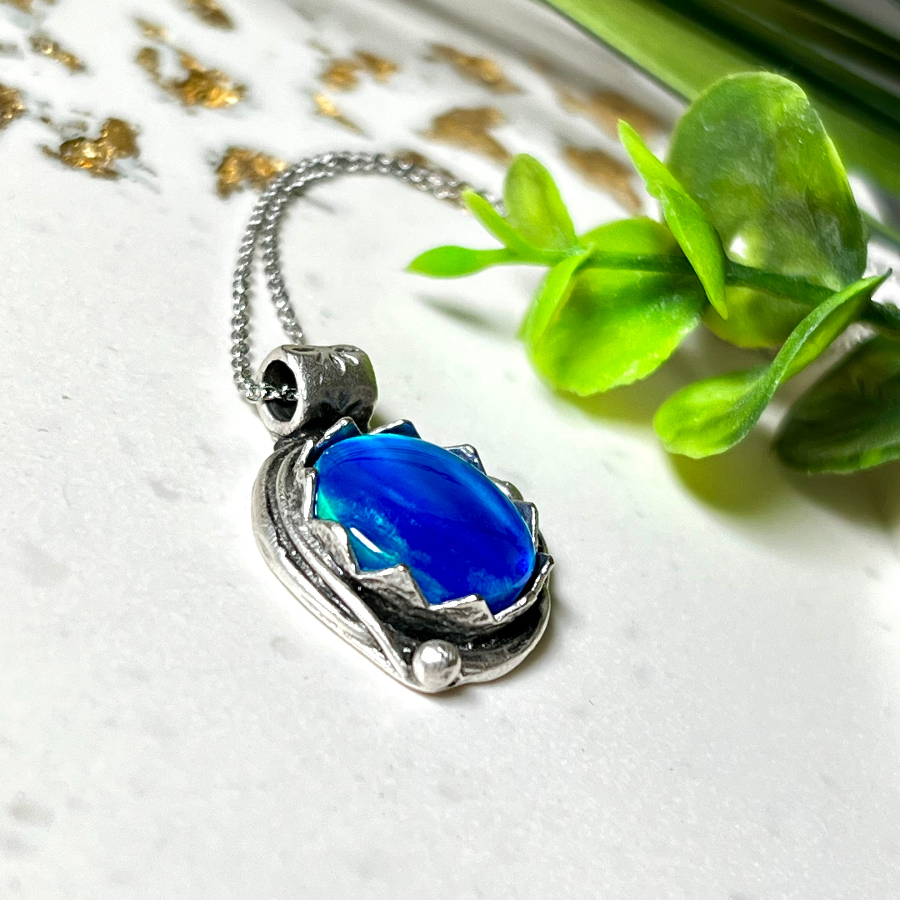 Handgemachter Anhänger mit rundem Cabochon aus Epoxidharz und schimmerndem synthetischem Opal. Die Farben changieren je nach Lichteinfall in Blau, Türkis und Grün. Der Opal sitzt in einer antik versilberten Fassung aus Messing (blei- & nickelfrei). Geliefert mit einer rhodinierten 925 Silberkette (45 cm). Jedes Stück ein Einzelstück – perfekt als stilvolles Geschenk oder persönlicher Glücksbringer.