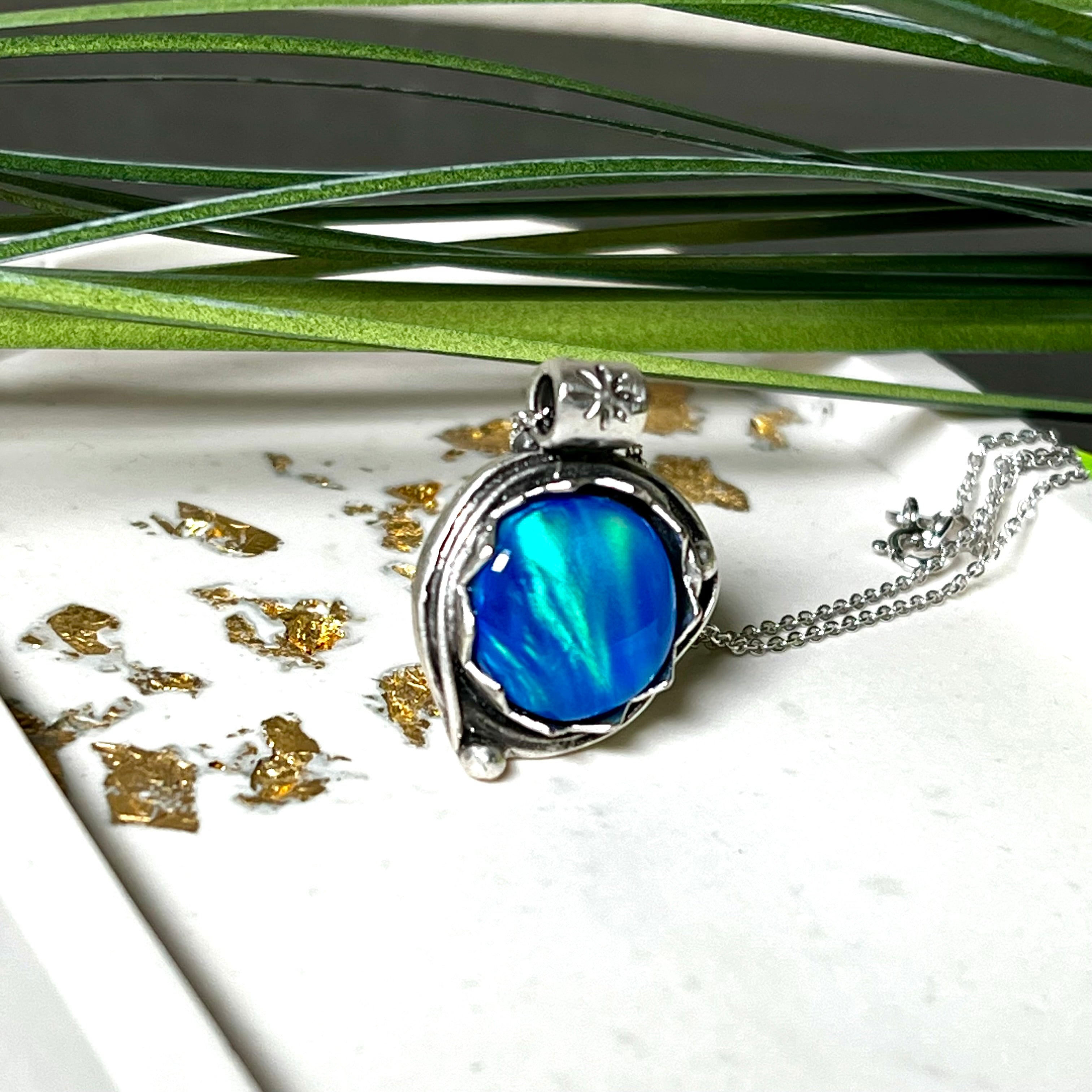 Handgemachter Anhänger mit rundem Cabochon aus Epoxidharz und schimmerndem synthetischem Opal. Die Farben changieren je nach Lichteinfall in Blau, Türkis und Grün. Der Opal sitzt in einer antik versilberten Fassung aus Messing (blei- & nickelfrei). Geliefert mit einer rhodinierten 925 Silberkette (45 cm). Jedes Stück ein Einzelstück – perfekt als stilvolles Geschenk oder persönlicher Glücksbringer.