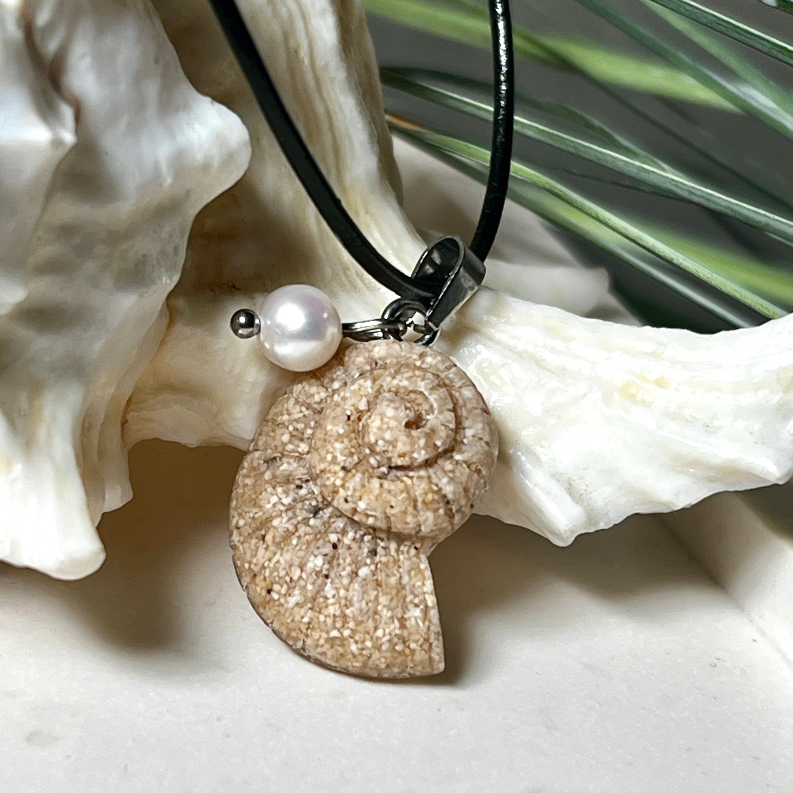 Handgefertigter Anhänger in Ammonit-Form aus echtem Meersand und Harz, kombiniert mit einer weißen Süßwasserperle. Der maritime Anhänger hängt an einer schwarzen Lederkette mit Edelstahlöse und Karabinerverschluss. Im Hintergrund liegt eine weiße Muschel, die das natürliche, strandinspirierte Design des Schmuckstücks unterstreicht. Ideal als besonderes Geschenk oder Erinnerung an den Sommer.