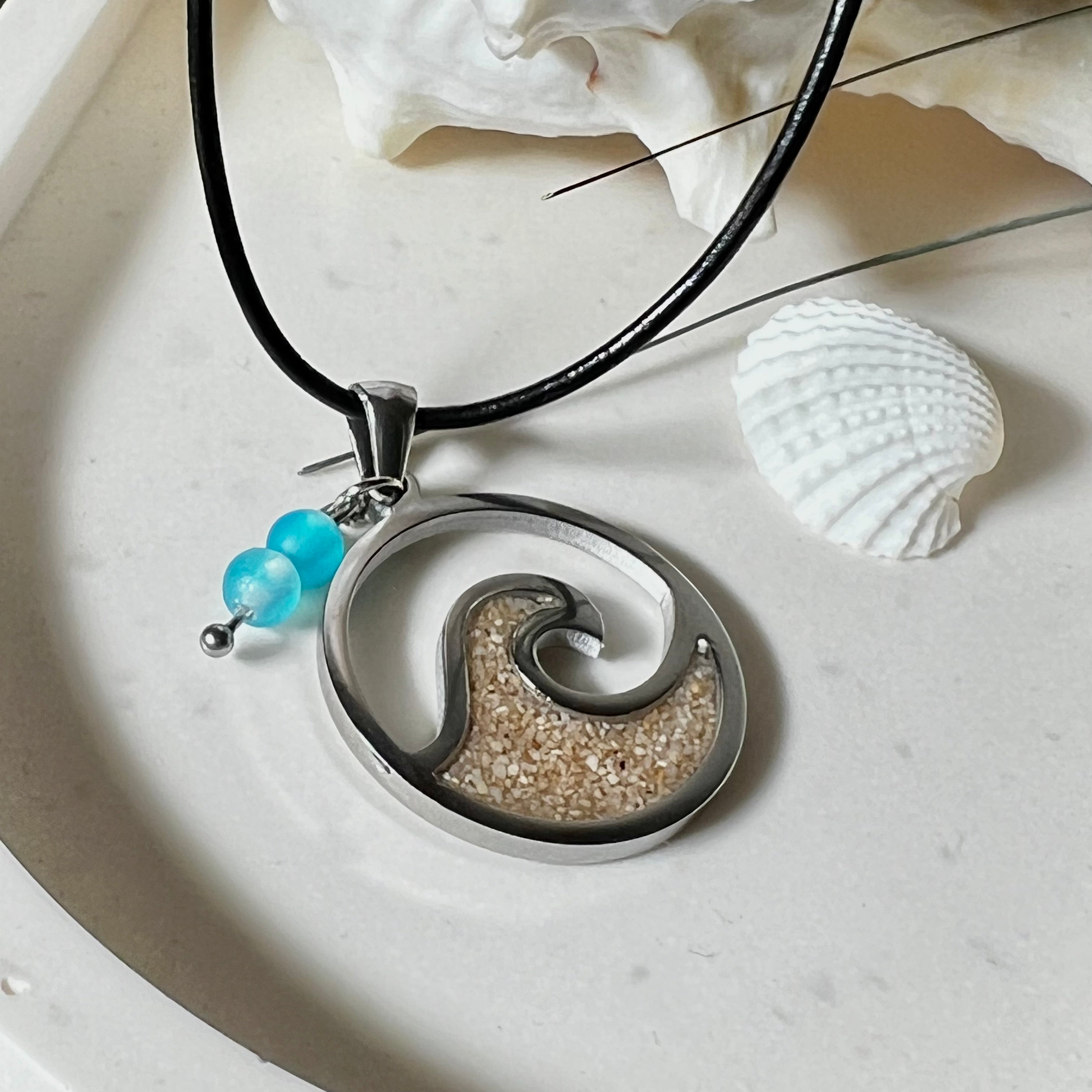 Wave Spirit – runder Edelstahlanhänger mit echter Sandfüllung in Harz eingebettet, inspiriert von einer stilisierten Welle. Verziert mit zwei blauen Achatperlen. Der Anhänger (ca. 2,5 cm Durchmesser) hängt an einer 45 cm langen Lederkette mit Edelstahlverschluss und 5 cm Verlängerungskette. Maritimer Look trifft moderne Eleganz – ein handgefertigtes Schmuckstück für Meerliebhaber