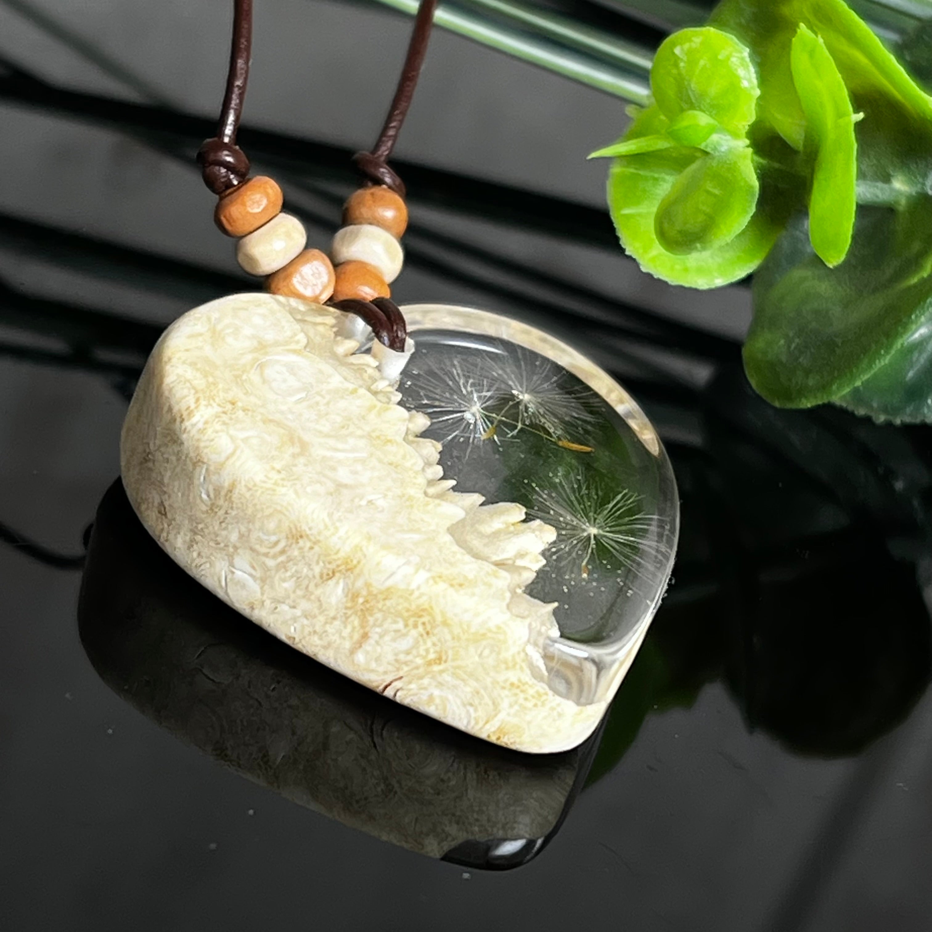 Herzanhänger aus Goldfield-Maserholz und transparentem Epoxidharz mit echter Pusteblume. Das handgefertigte Unikat kombiniert die natürliche Maserung des Holzes mit der zarten Leichtigkeit schwebender Pusteblumenschirmchen. Das braune Lederband mit Holzperlen ist verstellbar und verleiht dem Schmuckstück eine naturnahe, elegante Ausstrahlung.
