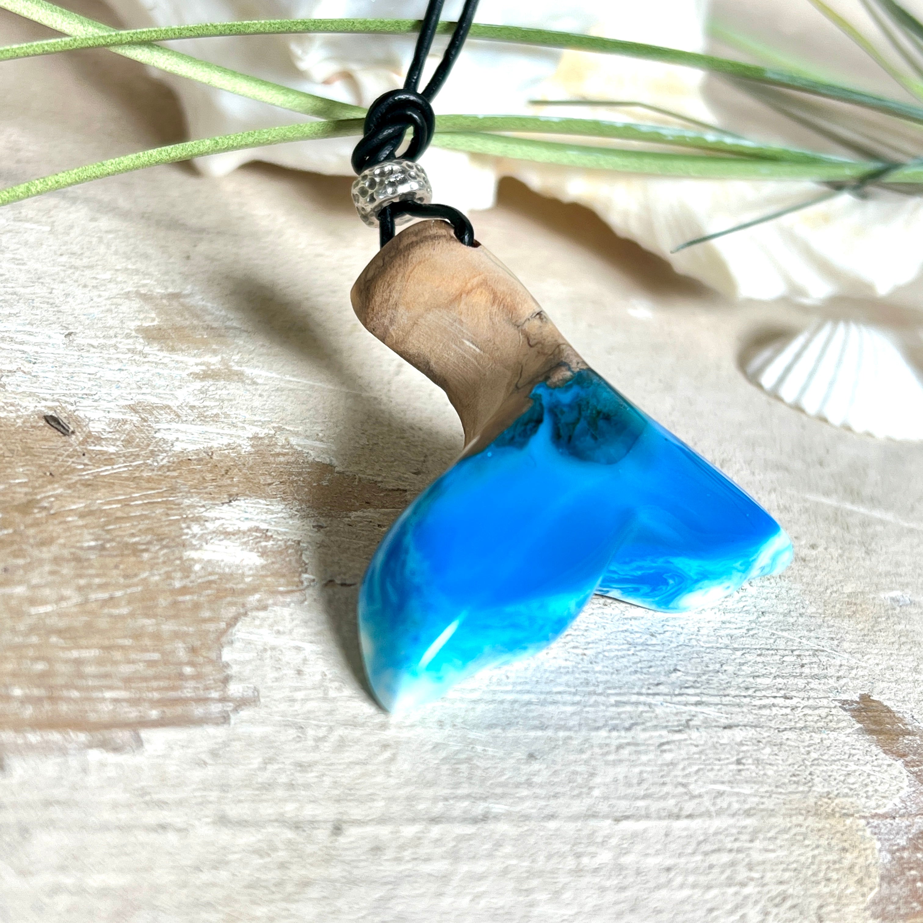 Handgefertigter Walflossen-Anhänger aus Olivenholz und glänzendem Harz in Blau-Grün-Weiß – maritimer Schmuck „Flossenklang“. Der Anhänger erinnert an die Form einer Walflosse und vereint die natürliche Maserung des Holzes mit dem schimmernden Farbspiel des Harzes. Mit verstellbarem schwarzen Lederband und Spacerperle. Ideal für Meeresliebhaber und Naturliebhaber – jedes Stück ein Unikat.