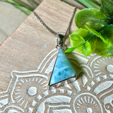 Larimar Anhänger in markanter Dreiecksform, eingefasst in rhodiniertes 925 Sterling Silber, vor strukturiertem Hintergrund mit grünen Blättern – stilvoller Edelsteinschmuck mit karibischem Flair.