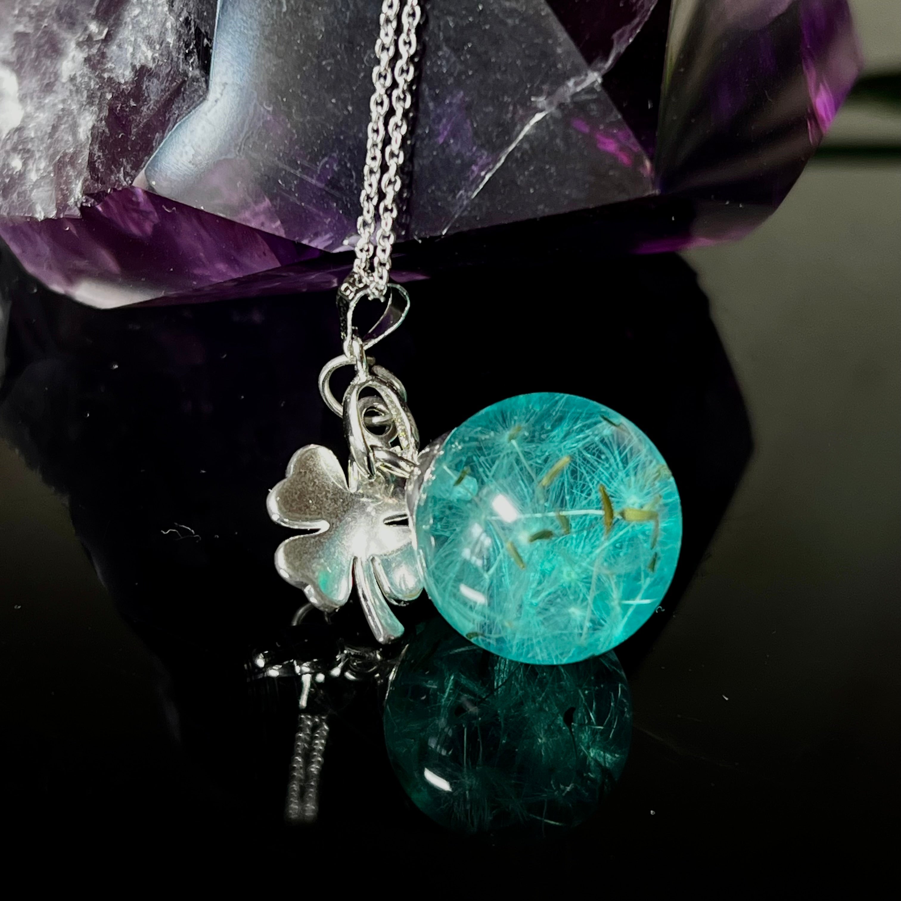 Eine handgefertigten Halskette mit türkiser Harzkugel, in der echte Pusteblumensamen eingeschlossen sind. Ergänzt durch einen glänzenden Edelstahl-Kleeblatt-Charm und einer feinen rhodinierten 925 Silberkette. Der Anhänger symbolisiert Glück, Leichtigkeit und neue Wege – ein emotionales Schmuckstück mit Bedeutung.