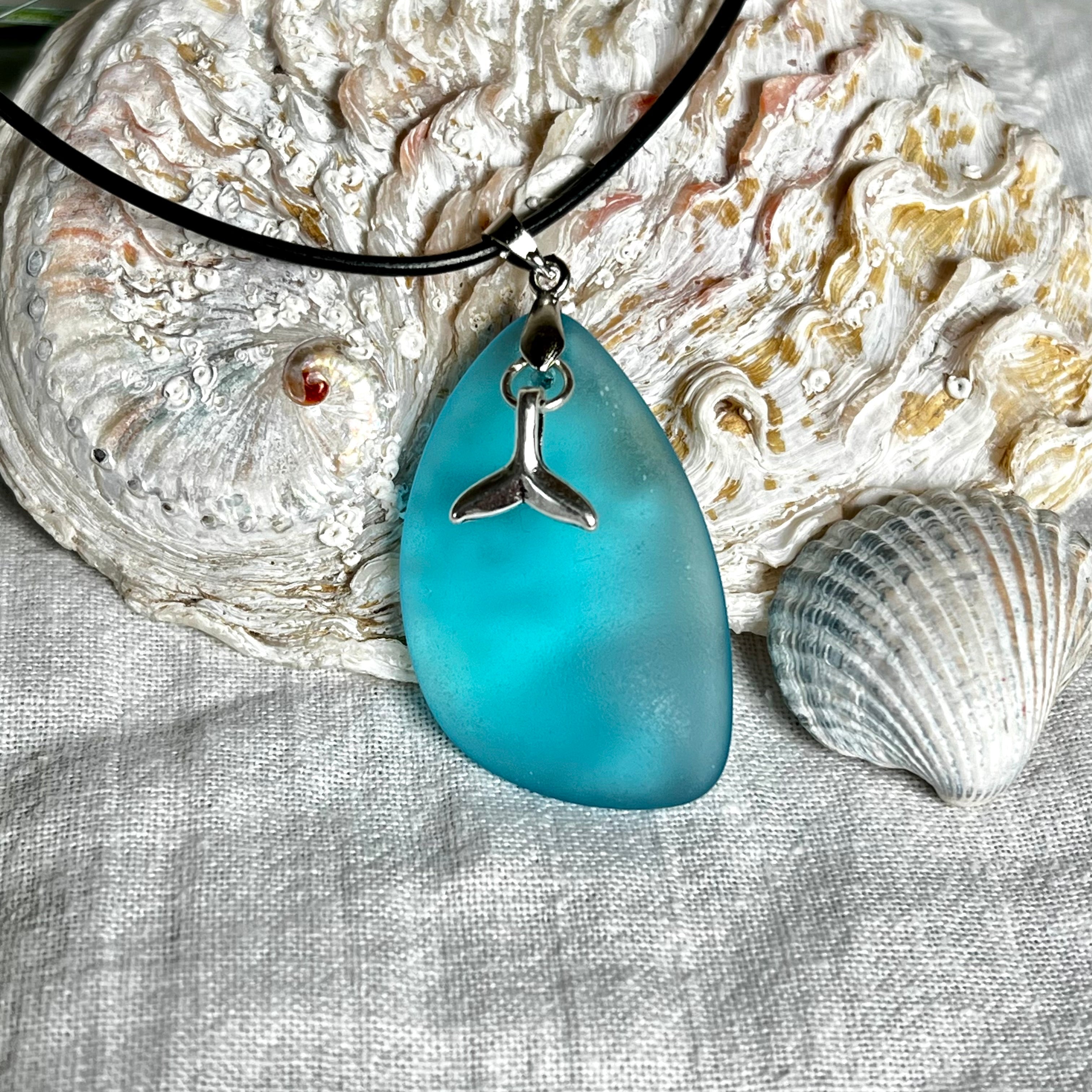 Edle Halskette „Blue Essence“ mit handgefertigtem türkisfarbenem Seeglas-Anhänger und Edelstahl-Flosse. Die Kette besteht aus schwarzer Lederkordel mit Karabinerverschluss und Verlängerung aus Edelstahl. Das matte Seeglas reflektiert Licht wie Meereswasser – ideal für Strandliebhaber und Naturliebhaber.