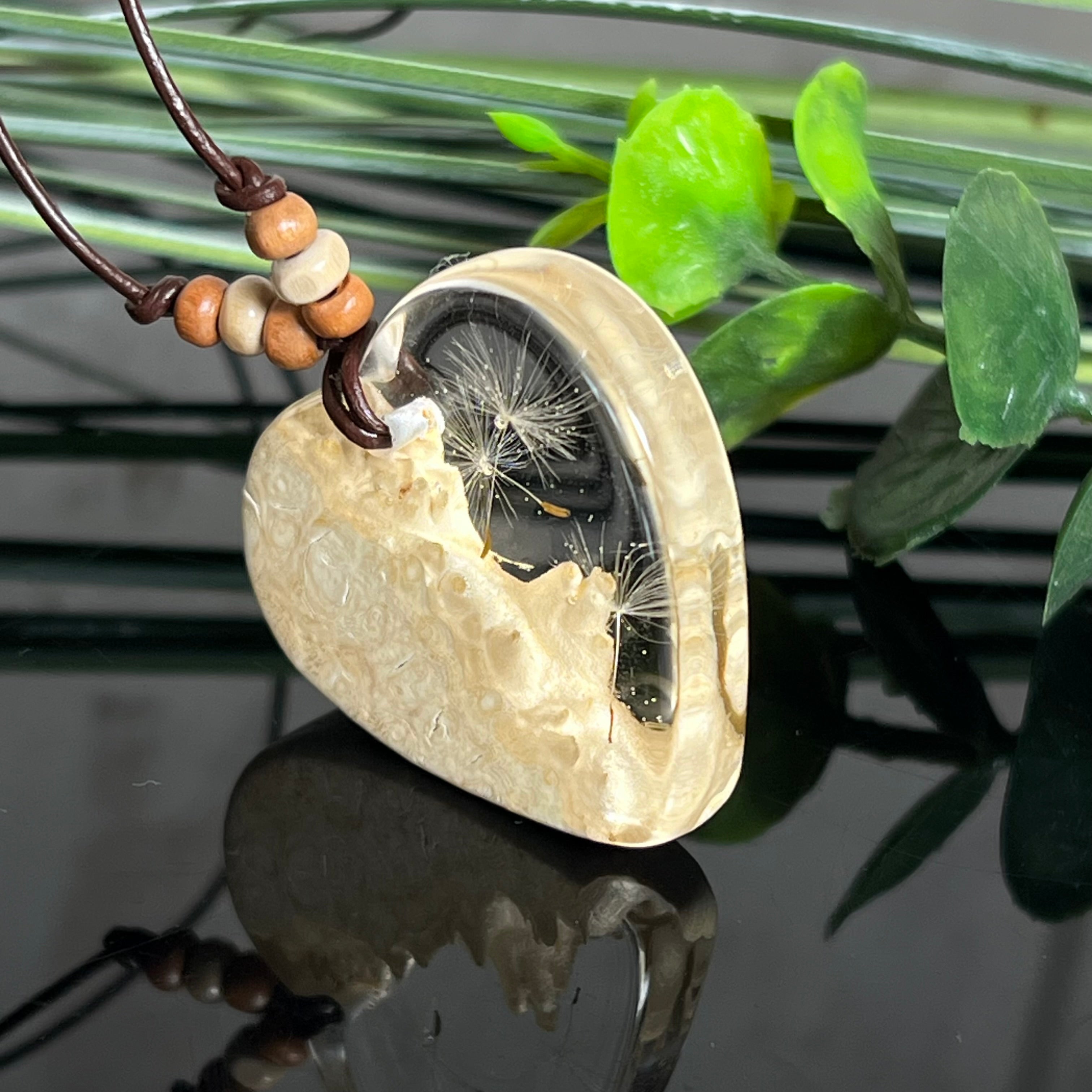 Herzanhänger aus Goldfield-Maserholz und transparentem Epoxidharz mit echter Pusteblume. Das handgefertigte Unikat kombiniert die natürliche Maserung des Holzes mit der zarten Leichtigkeit schwebender Pusteblumenschirmchen. Das braune Lederband mit Holzperlen ist verstellbar und verleiht dem Schmuckstück eine naturnahe, elegante Ausstrahlung.