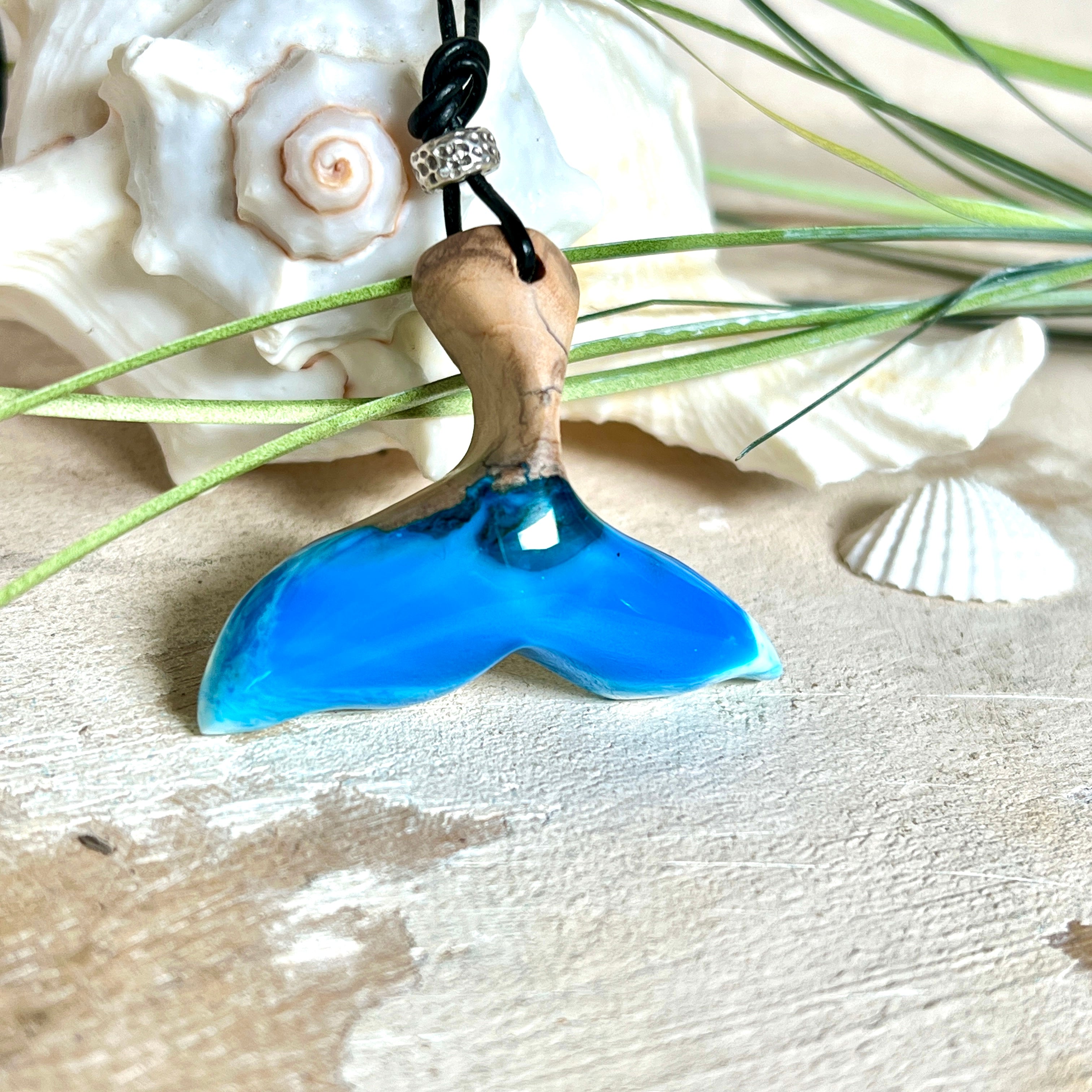 Handgefertigter Walflossen-Anhänger aus Olivenholz und glänzendem Harz in Blau-Grün-Weiß – maritimer Schmuck „Flossenklang“. Der Anhänger erinnert an die Form einer Walflosse und vereint die natürliche Maserung des Holzes mit dem schimmernden Farbspiel des Harzes. Mit verstellbarem schwarzen Lederband und Spacerperle. Ideal für Meeresliebhaber und Naturliebhaber – jedes Stück ein Unikat.