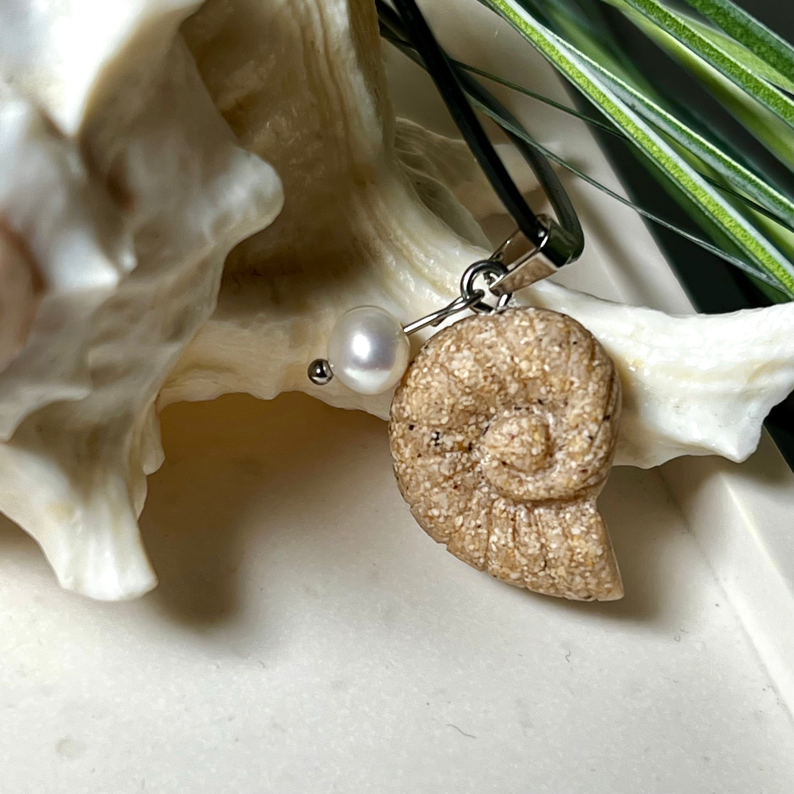 Handgefertigter Ammonit-Anhänger aus echtem Meersand & Harz mit filigraner Edelstahlöse und einer edlen Süßwasserperle. Maritimer Schmuck, inspiriert von der Kraft und Schönheit des Ozeans. Jedes Stück ein Unikat. Präsentiert auf einer Muschel, mit Lederkette.