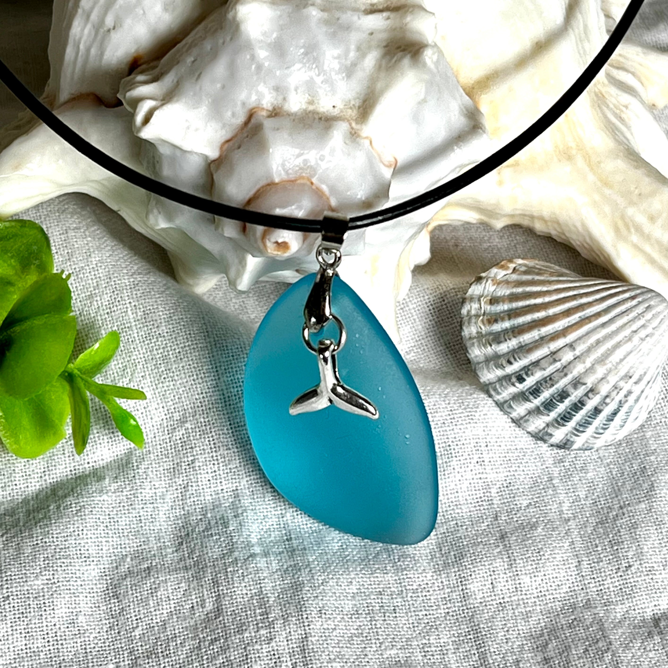 Edle Halskette „Blue Essence“ mit handgefertigtem türkisfarbenem Seeglas-Anhänger und Edelstahl-Flosse. Die Kette besteht aus schwarzer Lederkordel mit Karabinerverschluss und Verlängerung aus Edelstahl. Das matte Seeglas reflektiert Licht wie Meereswasser – ideal für Strandliebhaber und Naturliebhaber.