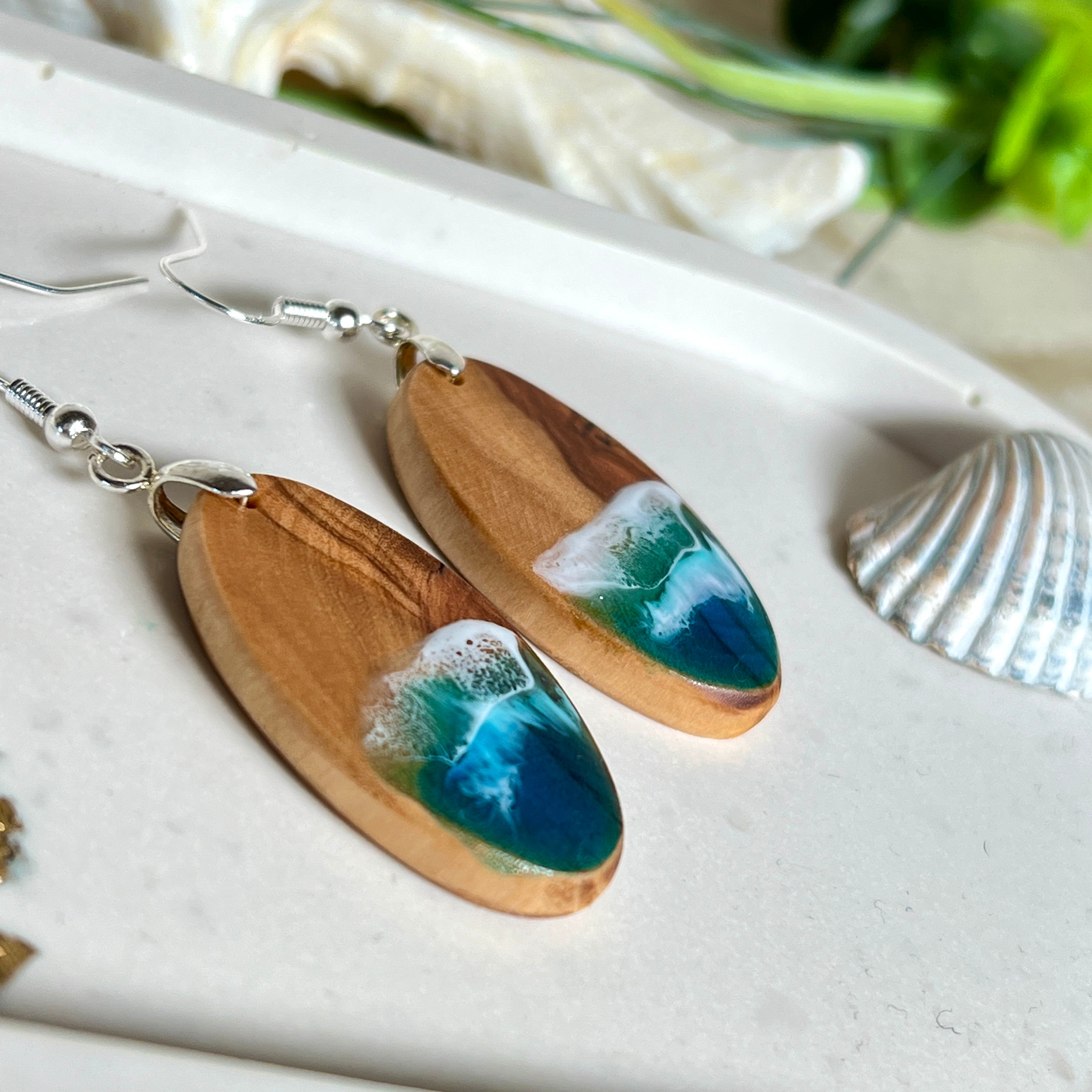 Handgefertigte Ohrringe „Wave Harmony“ aus Olivenholz und Epoxidharz in Blau-, Türkis- und Weißtönen. Das Design erinnert an Meereswellen am Strand. Jeder Ohrring ist ein Unikat mit individueller Maserung. Versilberte, nickelfreie und bleifreie Anhängerschlaufe, 925 Silber Ohrhaken. Maritimer Schmuck – perfekt für Meerliebhaber und als edles Geschenk.