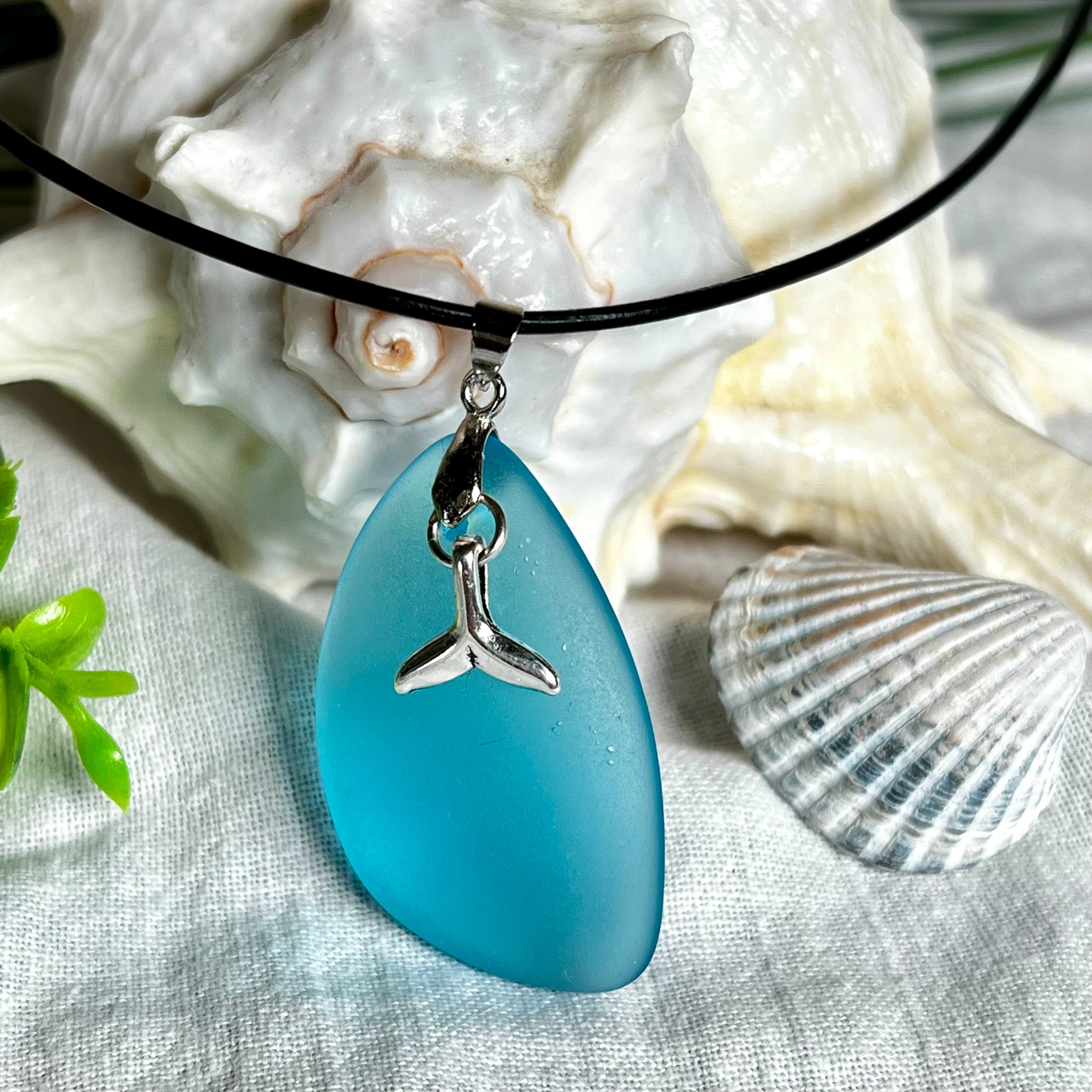 Edle Halskette „Blue Essence“ mit handgefertigtem türkisfarbenem Seeglas-Anhänger und Edelstahl-Flosse. Die Kette besteht aus schwarzer Lederkordel mit Karabinerverschluss und Verlängerung aus Edelstahl. Das matte Seeglas reflektiert Licht wie Meereswasser – ideal für Strandliebhaber und Naturliebhaber.