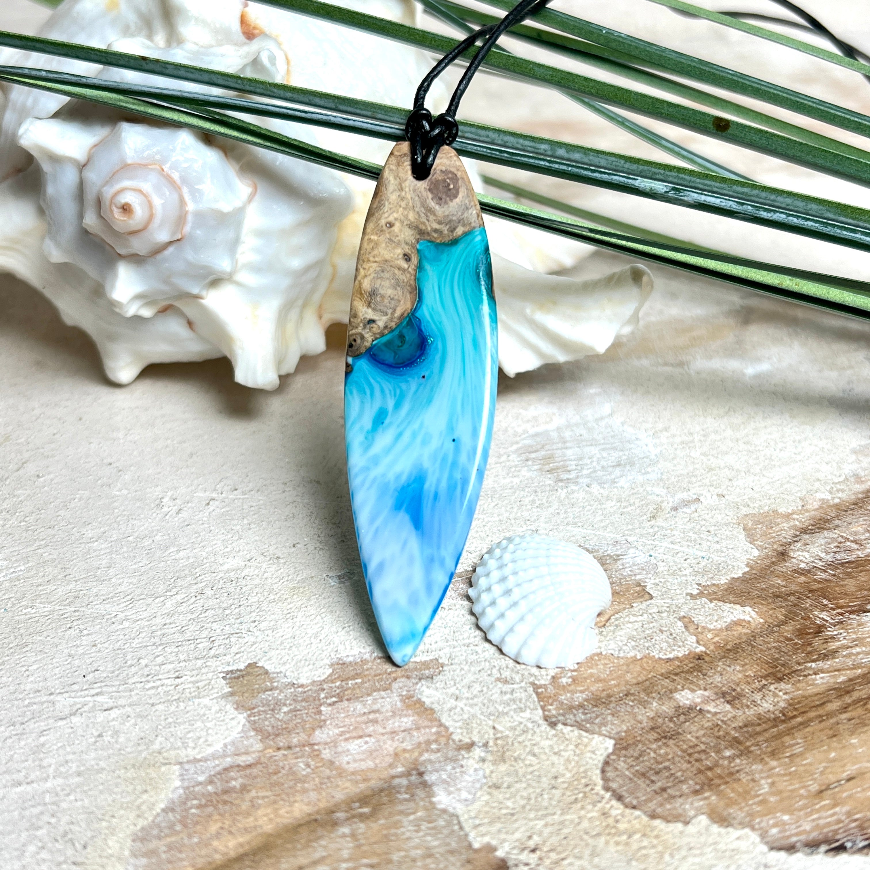 Surfbrett-Anhänger handgefertigt aus Maserholz & Harz in Blau-Weiß. Größe ca. 6,3 × 2 cm. Lederband verstellbar 26–47 cm, Schiebeknoten mit Holzperlen.