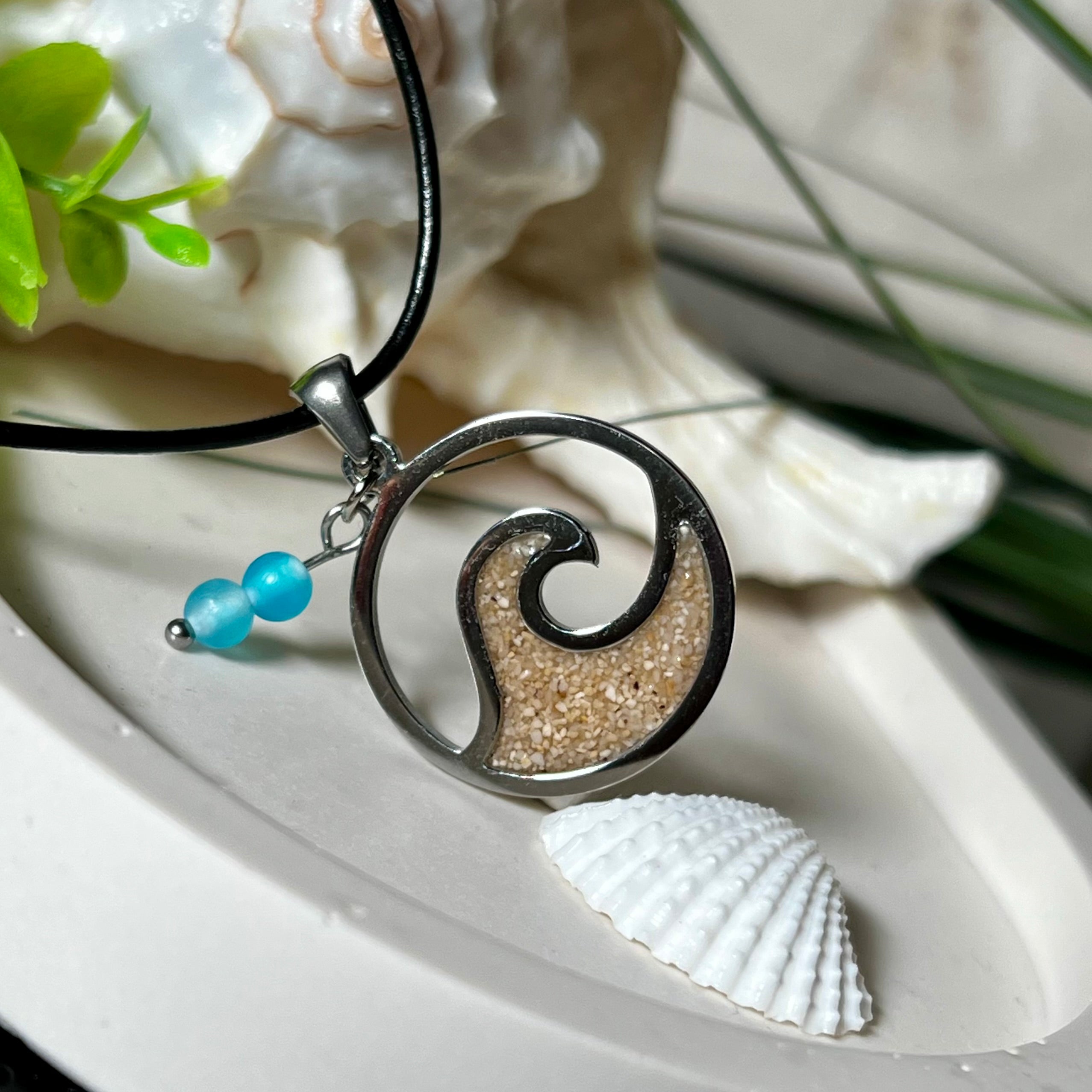 Wave Spirit – runder Edelstahlanhänger mit echter Sandfüllung in Harz eingebettet, inspiriert von einer stilisierten Welle. Verziert mit zwei blauen Achatperlen. Der Anhänger (ca. 2,5 cm Durchmesser) hängt an einer 45 cm langen Lederkette mit Edelstahlverschluss und 5 cm Verlängerungskette. Maritimer Look trifft moderne Eleganz – ein handgefertigtes Schmuckstück für Meerliebhaber