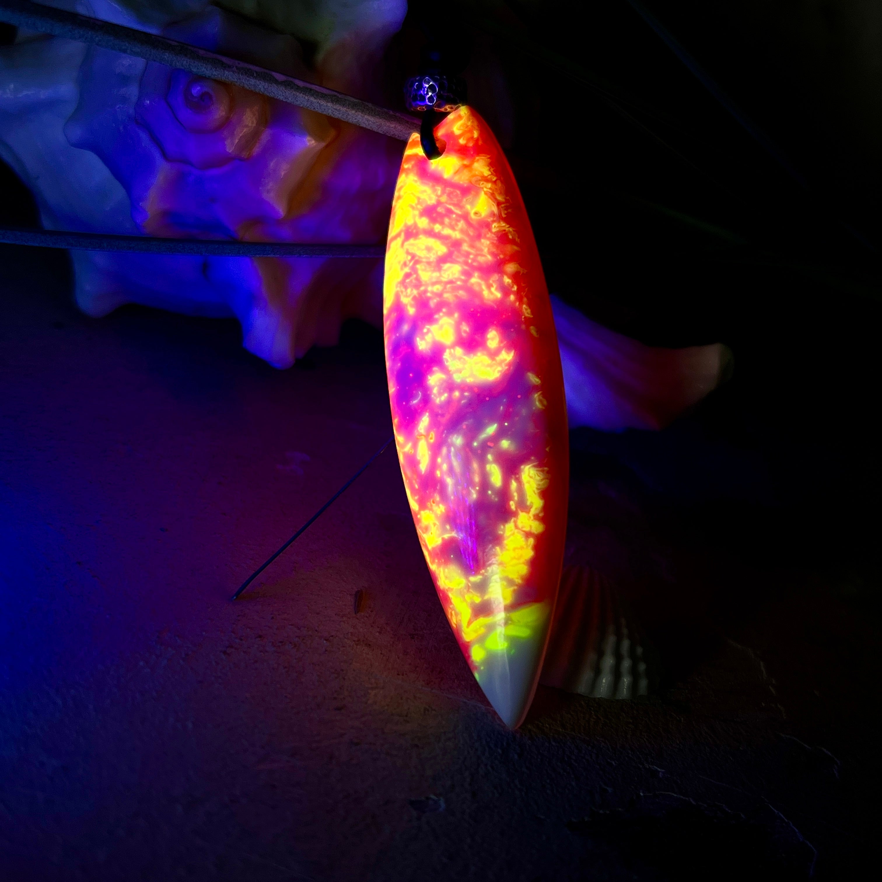 Handgefertigter Surfbrett-Anhänger aus fluoreszierendem Epoxidharz in leuchtenden Orange-, Rot- und Gelbtönen. Leuchtet bei Schwarzlicht. Kombiniert mit verstellbarem Lederband (26–45 cm), verziert mit Spacer- und Holzperlen. Das Design ist inspiriert von der Farbintensität eines Sonnenuntergangs über dem Meer.