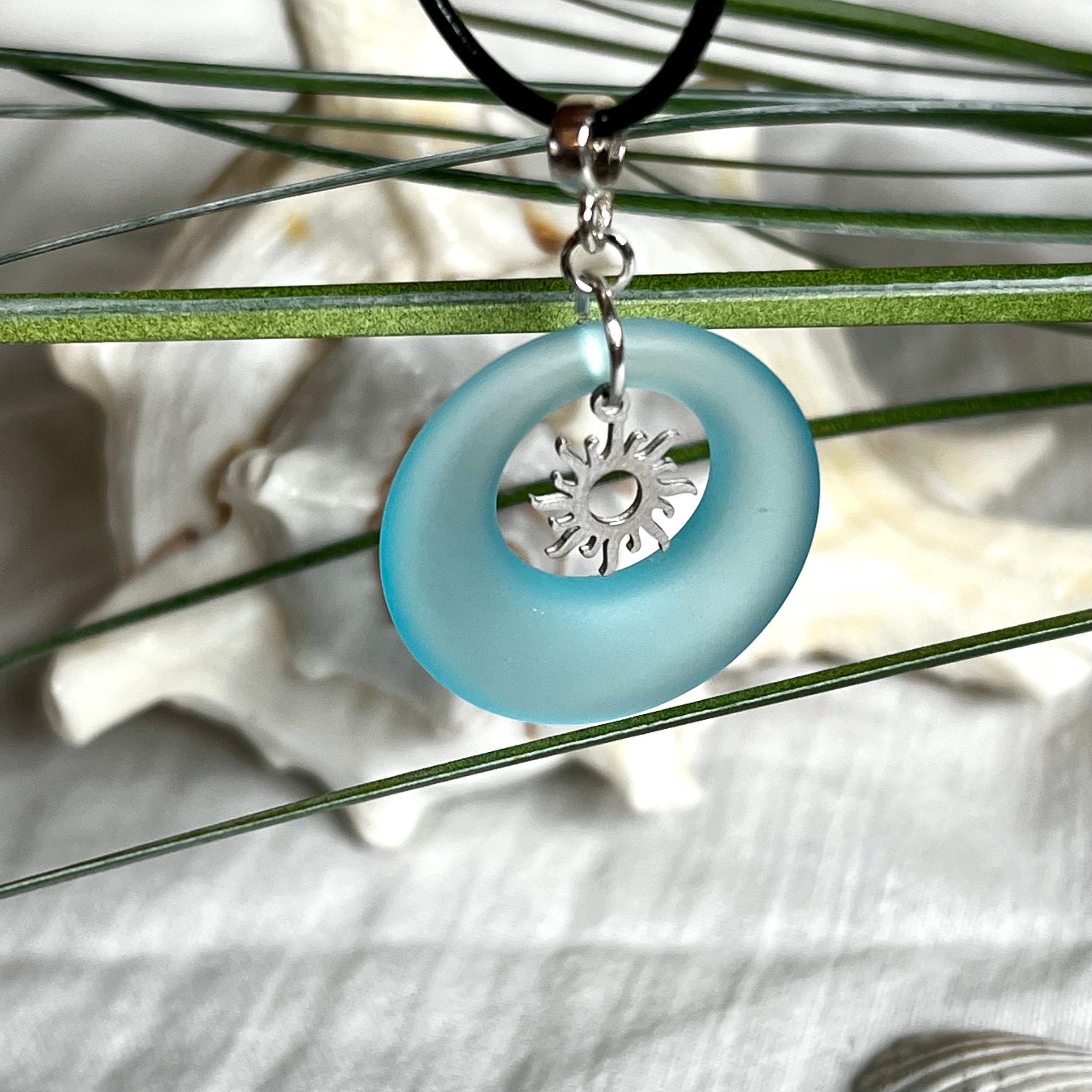 Handgefertigte Halskette mit Donut-Anhänger aus türkisfarbenem Seeglas, kombiniert mit einem filigranen Sonnen-Charm aus Edelstahl. Die Kette besteht aus einer schwarzen Lederschnur (1,5 mm) mit Karabiner und Verlängerungskette aus Edelstahl. Maritimes Schmuckstück für Meerliebhaberinnen und Sommerfreunde. Unikat mit stilvoller Ausstrahlung.