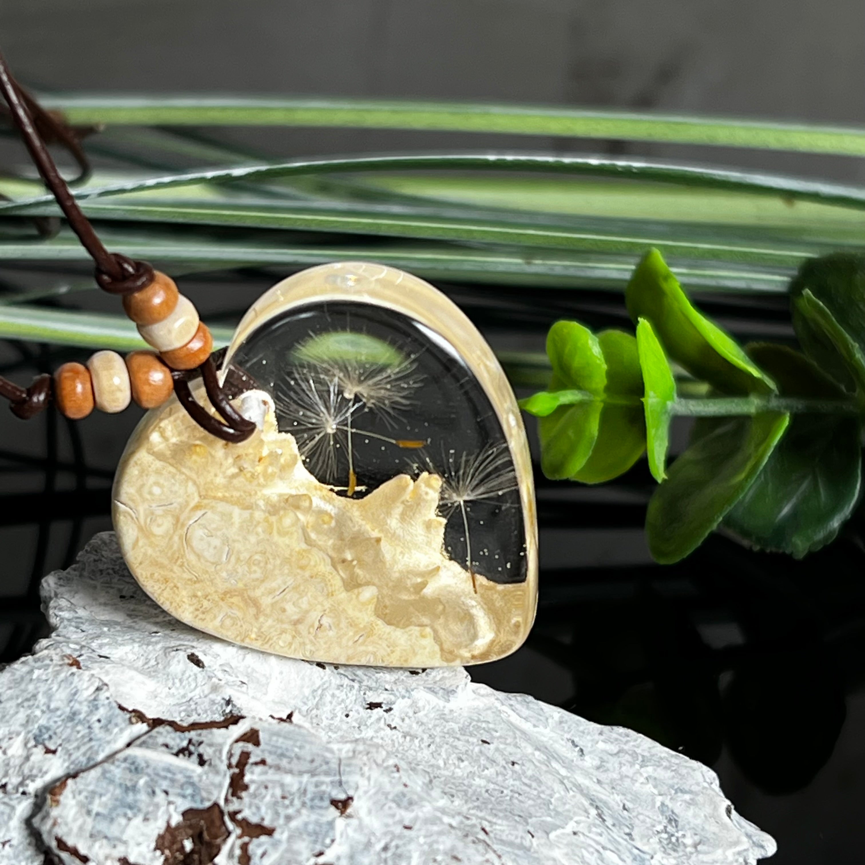 Herzanhänger aus Goldfield-Maserholz und transparentem Epoxidharz mit echter Pusteblume. Das handgefertigte Unikat kombiniert die natürliche Maserung des Holzes mit der zarten Leichtigkeit schwebender Pusteblumenschirmchen. Das braune Lederband mit Holzperlen ist verstellbar und verleiht dem Schmuckstück eine naturnahe, elegante Ausstrahlung.