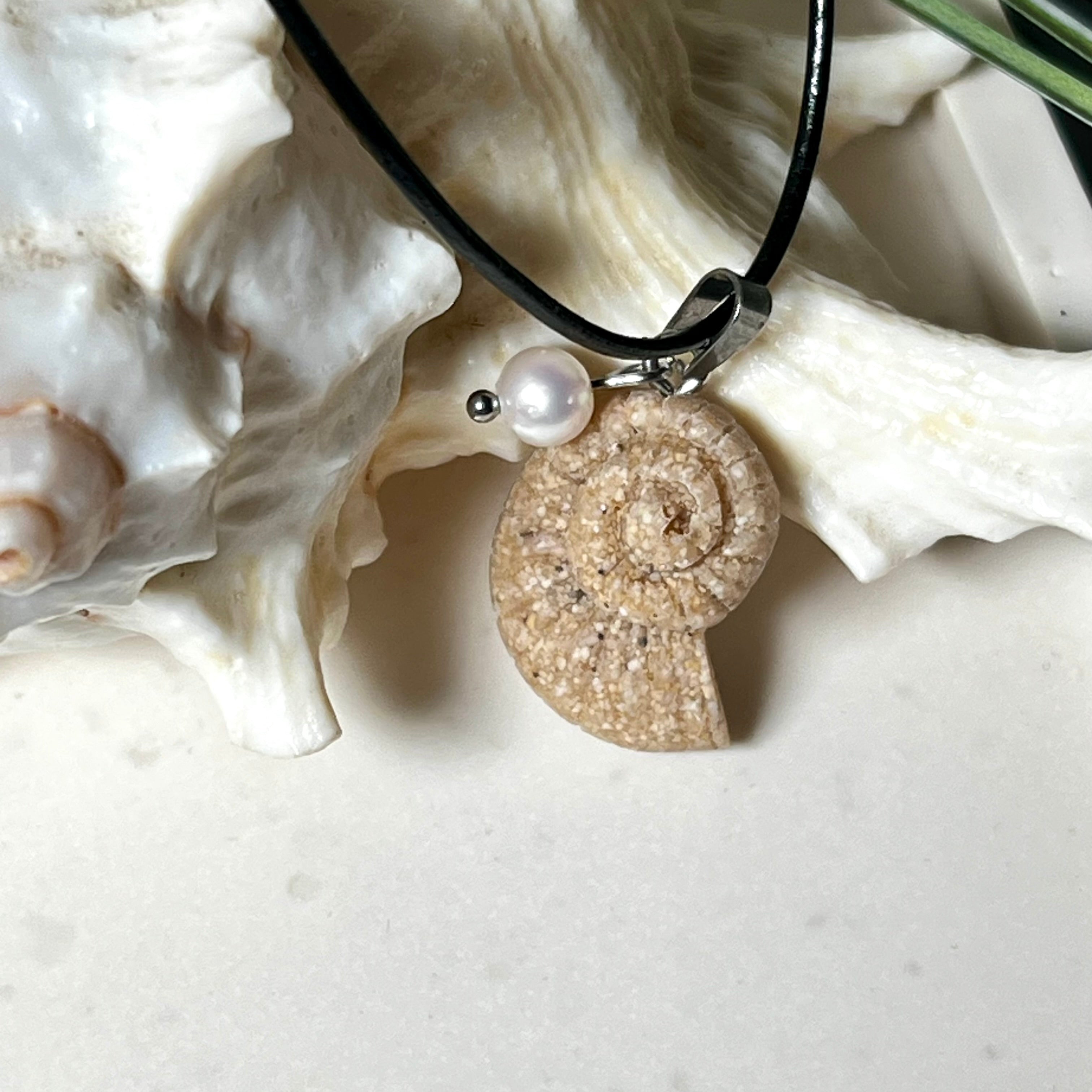 Handgefertigter Anhänger in Ammonit-Form aus echtem Meersand und Harz, kombiniert mit einer weißen Süßwasserperle. Der maritime Anhänger hängt an einer schwarzen Lederkette mit Edelstahlöse und Karabinerverschluss. Im Hintergrund liegt eine weiße Muschel, die das natürliche, strandinspirierte Design des Schmuckstücks unterstreicht. Ideal als besonderes Geschenk oder Erinnerung an den Sommer.