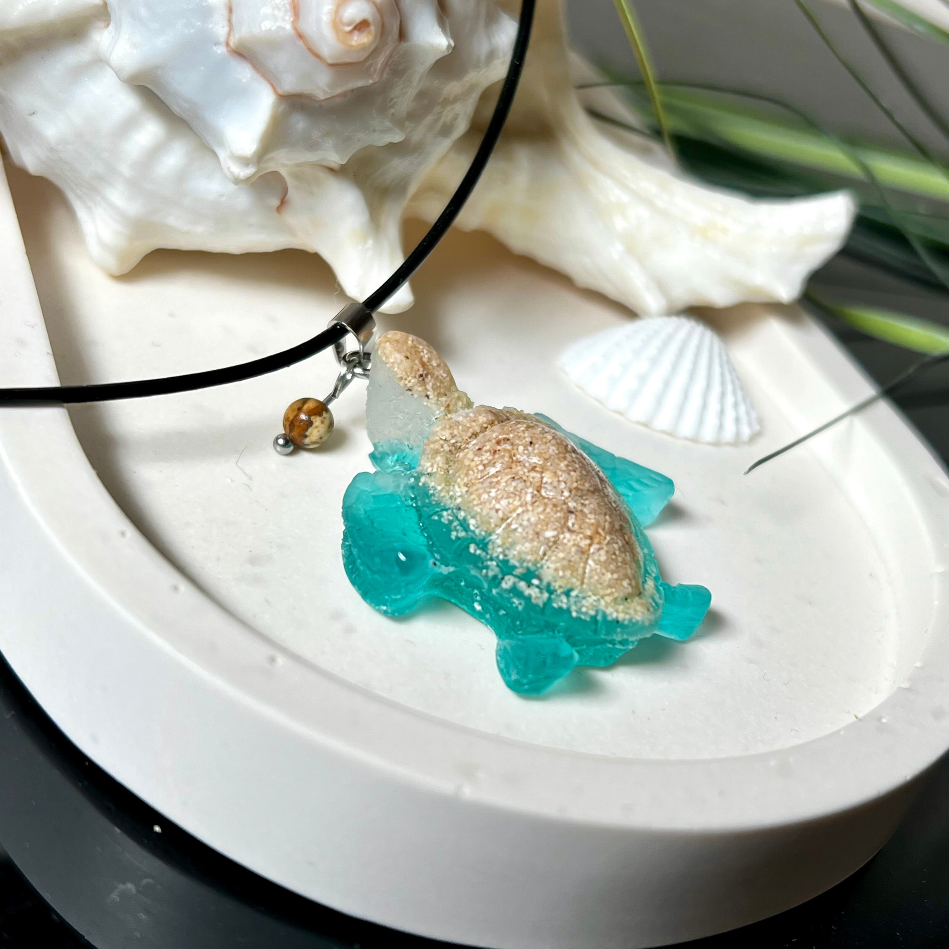 Handgefertigter Schildkröten-Anhänger Turtle Harmony aus echtem Meersand und transparent-türkisfarbenem Harz, kombiniert mit einer kleinen Jaspisperle. Symbol für Weisheit, Schutz und Harmonie. Mit Edelstahl-Anhängerschlaufe und Lederkette (45 + 5 cm). Maritimer Schmuck als Unikat, ideal als liebevolles Geschenk oder stilvolles Accessoire für den Alltag.