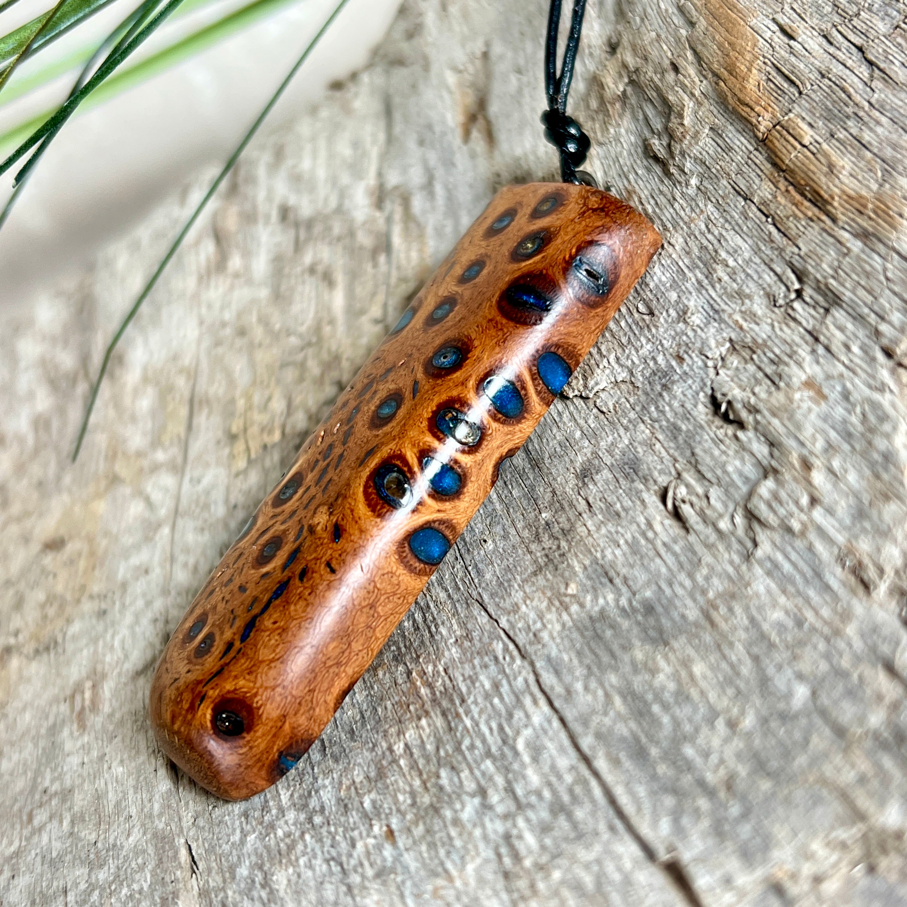 Handgefertigter Anhänger aus australischem Banksiazapfen und gegossenem Harz in Blau. Der rechteckige Anhänger (5,5 x 2 cm) zeigt eine charakteristische Maserung mit markanten Löchern, die durch die Samenstruktur des Zapfens entstehen. Das Lederband ist verstellbar (29–51 cm) und mit Holzperlen verziert. Ein edles Natur-Unikat.