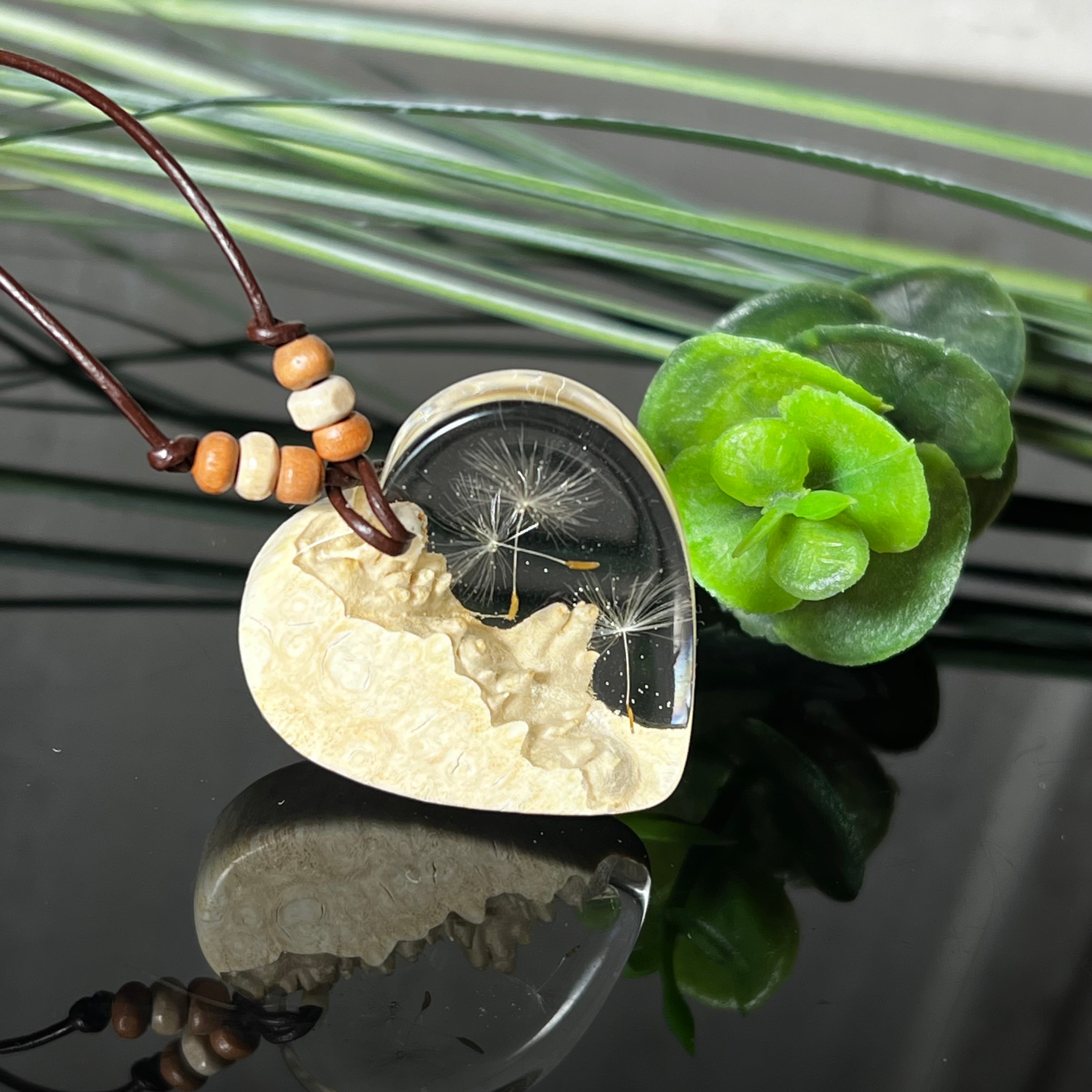Herzanhänger aus Goldfield-Maserholz und transparentem Epoxidharz mit echter Pusteblume. Das handgefertigte Unikat kombiniert die natürliche Maserung des Holzes mit der zarten Leichtigkeit schwebender Pusteblumenschirmchen. Das braune Lederband mit Holzperlen ist verstellbar und verleiht dem Schmuckstück eine naturnahe, elegante Ausstrahlung.