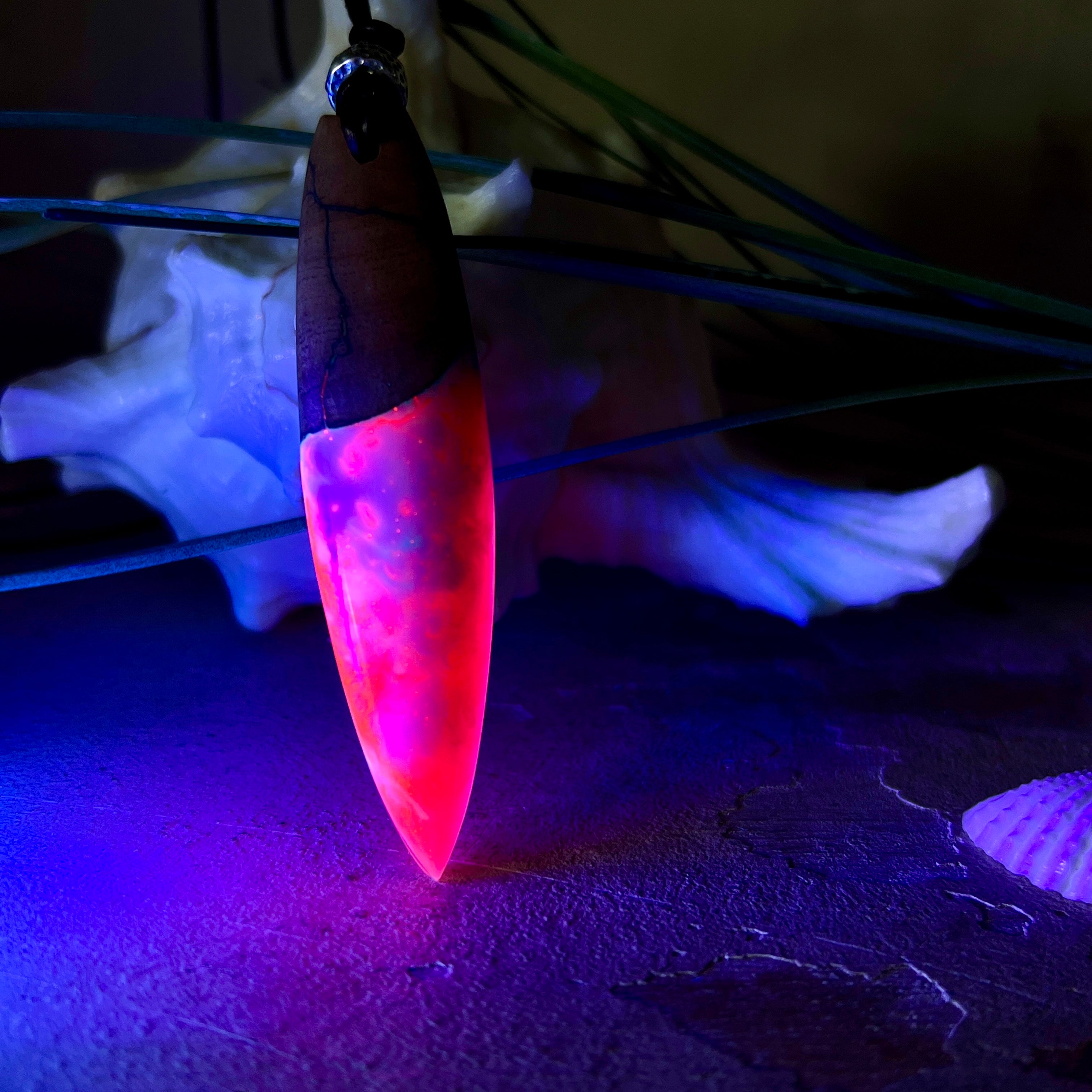 Handgefertigter Surfbrett-Anhänger aus Nussbaumholz und fluoreszierendem Epoxidharz in Pink, Blau und Weiß. Der Anhänger leuchtet zart bei Schwarzlicht und wird an einem verstellbaren Lederband (25–44 cm) mit Holzperlen getragen. Ein maritimes Unikat – ideal für Surfer und Naturliebhaber.