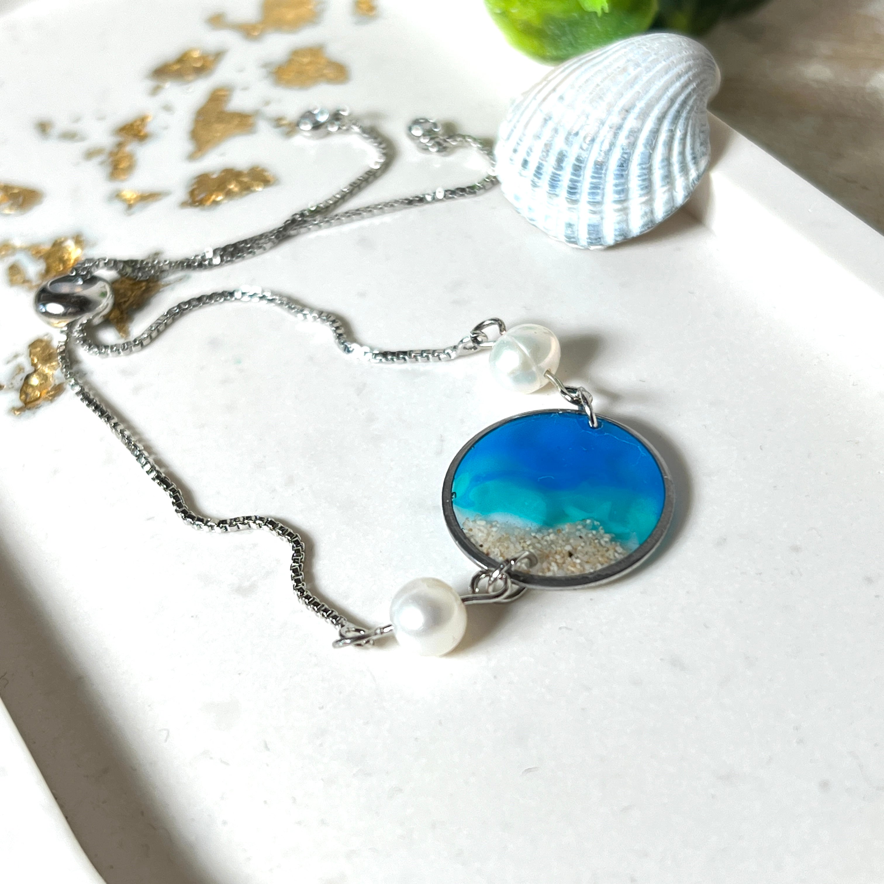 Elegantes Edelstahl-Armband mit rundem Resin-Anhänger in Meeresblau. Im Anhänger liegt echter Meersand als Ufersaum; darüber Sticker-Motive Seestern & Muschel. Zwei feine Süßwasserperlen verzieren das Armband. Verstellbarer Schiebeverschluss . Handgefertigtes Unikat – maritimer Schmuck, der an Wellenrauschen und Strandspaziergänge erinnert.