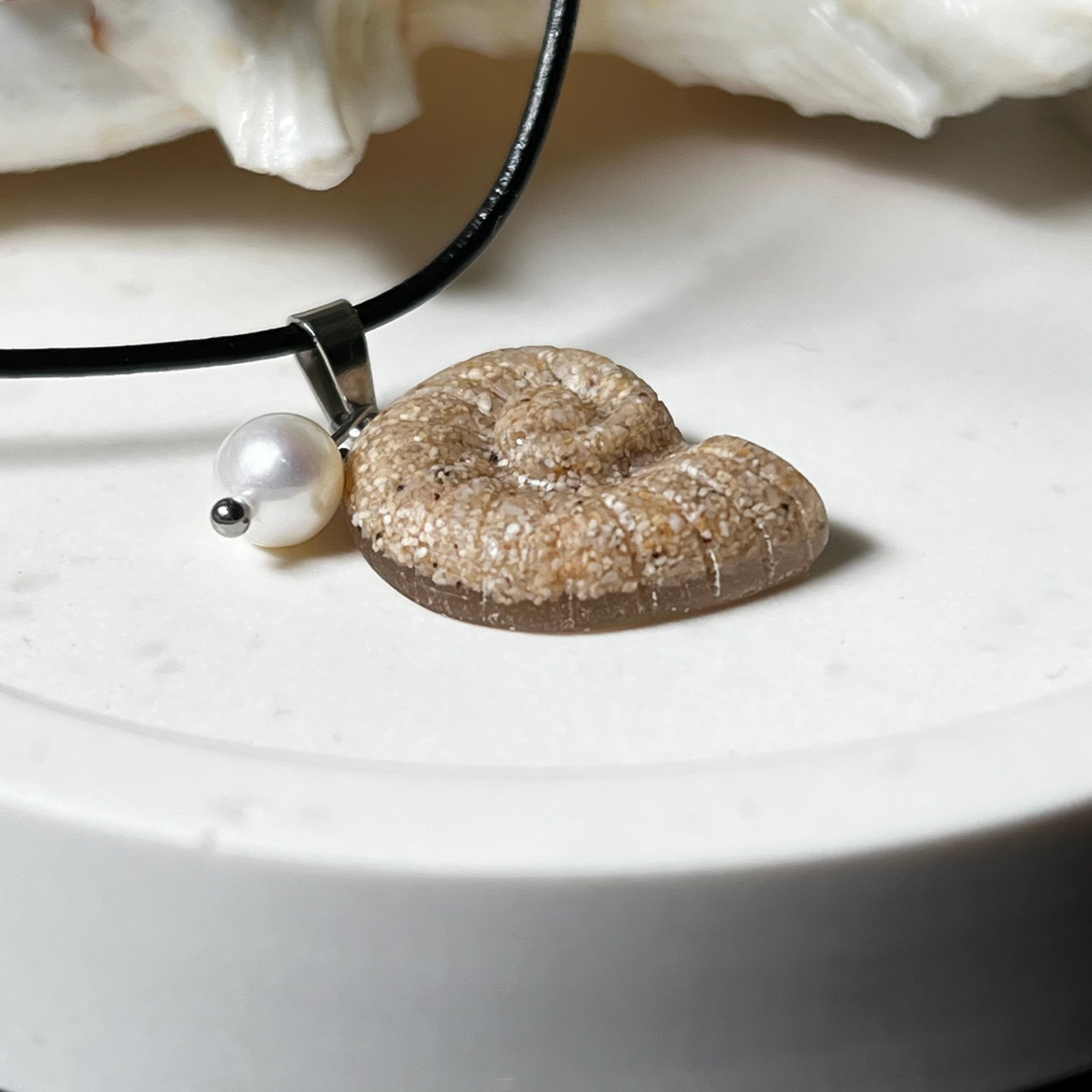 Handgefertigter Ammonit-Anhänger aus echtem Meersand & Harz mit filigraner Edelstahlöse und einer edlen Süßwasserperle. Maritimer Schmuck, inspiriert von der Kraft und Schönheit des Ozeans. Jedes Stück ein Unikat. Präsentiert auf einer Muschel, mit Lederkette.