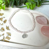 Handgefertigtes Rosenquarz-Armband mit facettierten 4mm Perlen, ergänzt durch einen filigranen Blume-des-Lebens-Anhänger aus 925er Silber. Präsentiert auf hellem Hintergrund mit Rosenquarzsteinen und goldenen Dekoelementen. Spiritueller Schmuck mit harmonisierender Wirkung.