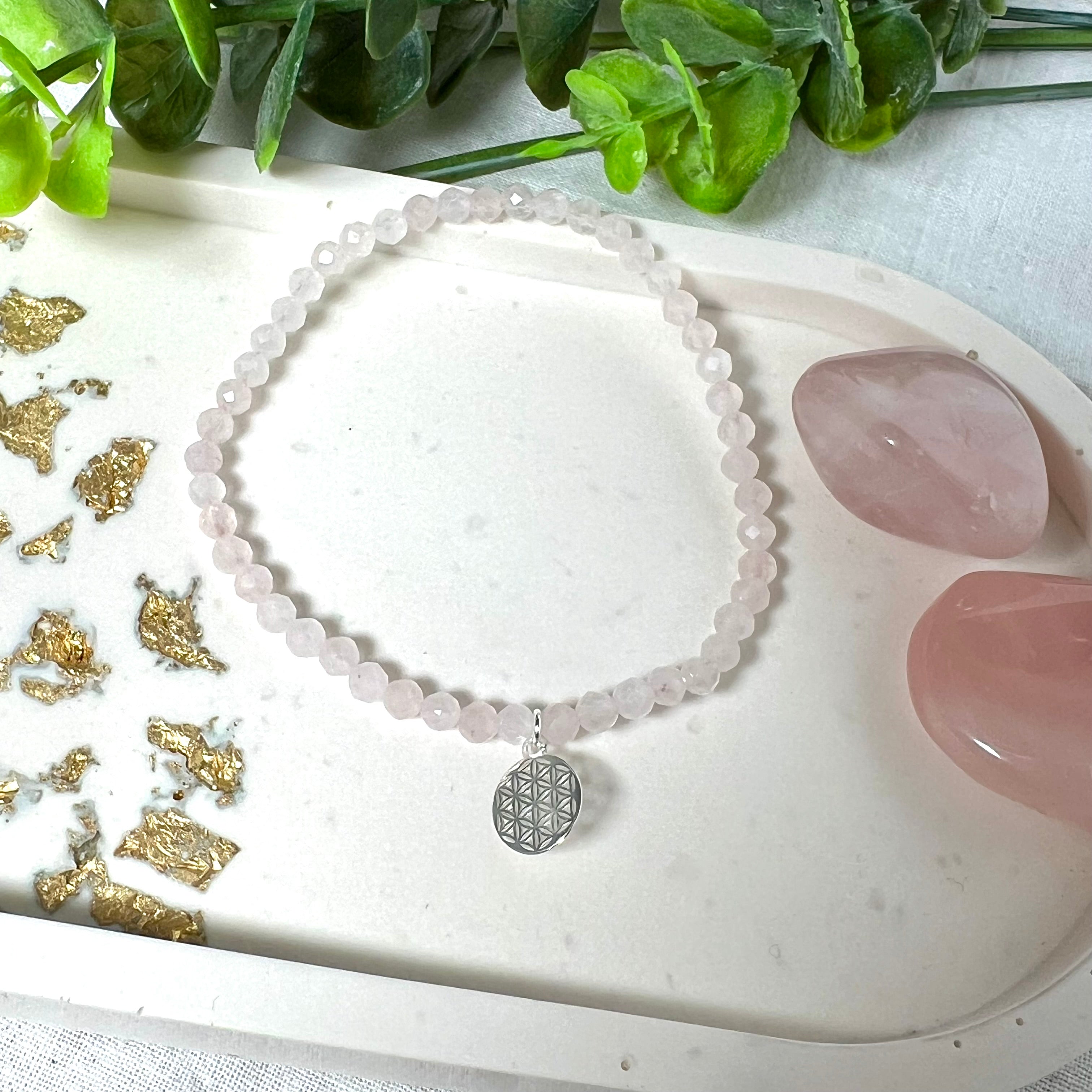 Handgefertigtes Rosenquarz-Armband mit facettierten 4mm Perlen, ergänzt durch einen filigranen Blume-des-Lebens-Anhänger aus 925er Silber. Präsentiert auf hellem Hintergrund mit Rosenquarzsteinen und goldenen Dekoelementen. Spiritueller Schmuck mit harmonisierender Wirkung.