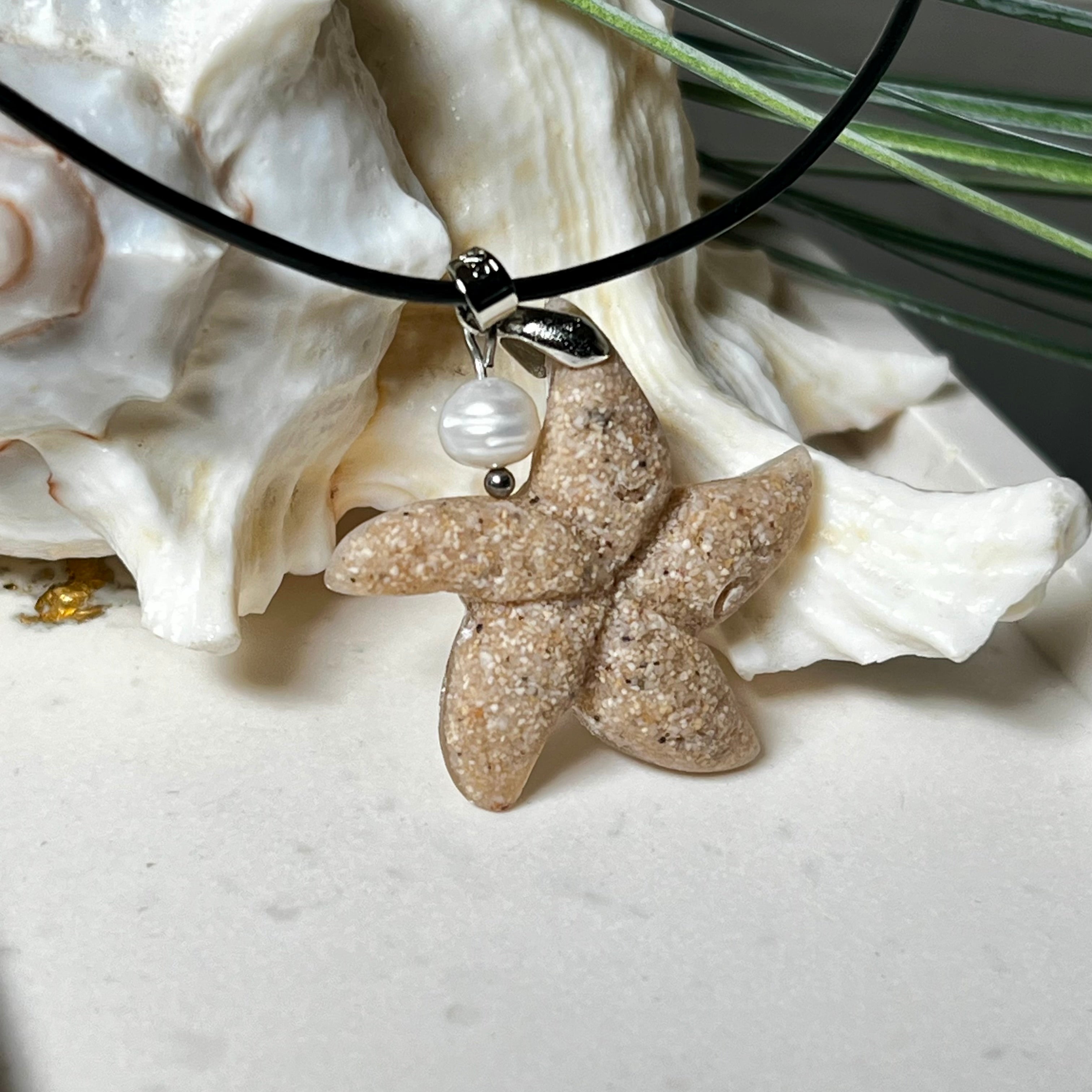 Handgefertigter Anhänger in Seesternform aus echtem Meersand und transparentem Harz. Verziert mit einer natürlichen Süßwasserperle. Edelstahlöse, Lederkette mit Karabinerverschluss (45 cm + 5 cm Verlängerung). Maritimer Schmuck im Unikatcharakter – inspiriert vom Meer und handgefertigt mit Liebe zum Detail. Perfektes Geschenk für Strandliebhaber.