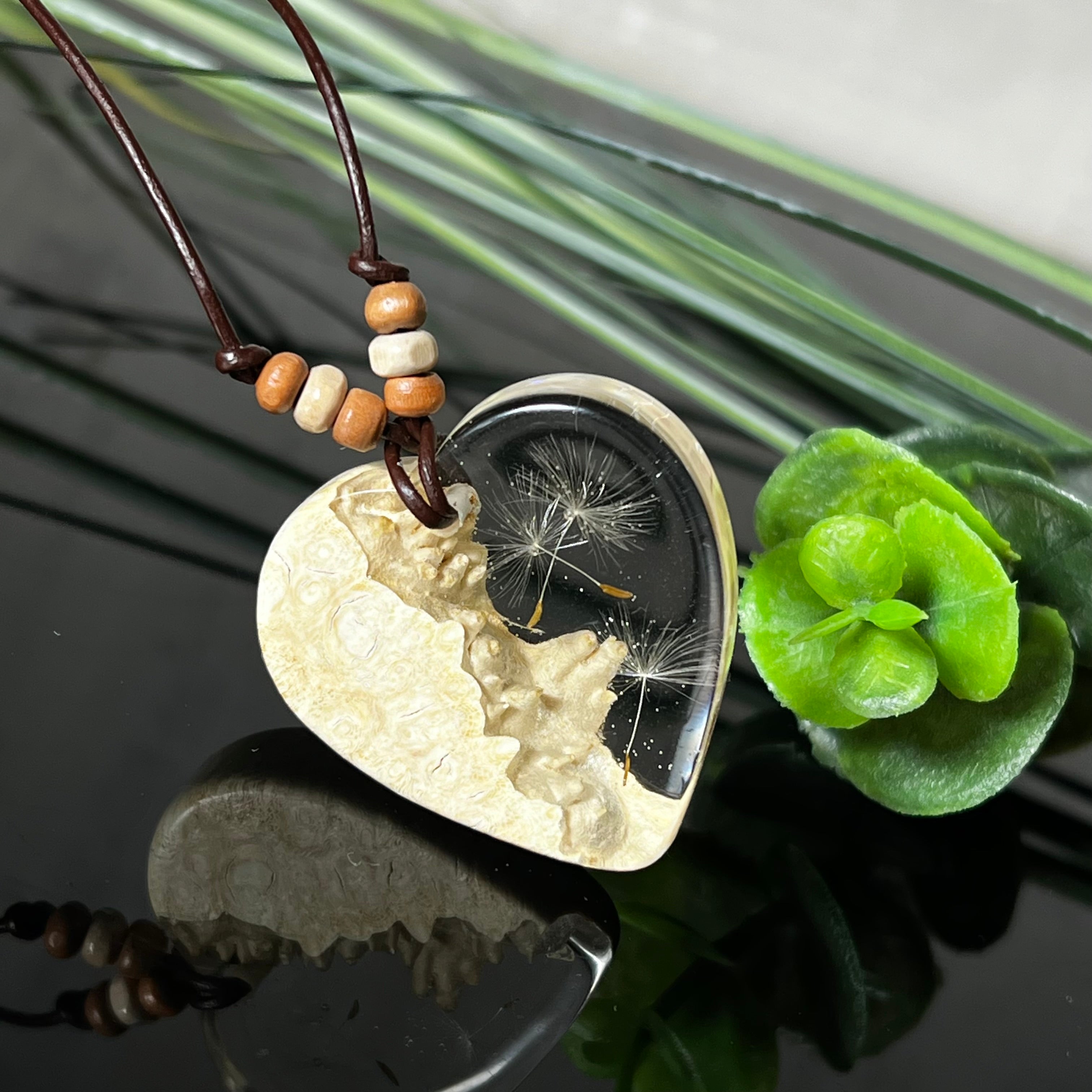 Herzanhänger aus Goldfield-Maserholz und transparentem Epoxidharz mit echter Pusteblume. Das handgefertigte Unikat kombiniert die natürliche Maserung des Holzes mit der zarten Leichtigkeit schwebender Pusteblumenschirmchen. Das braune Lederband mit Holzperlen ist verstellbar und verleiht dem Schmuckstück eine naturnahe, elegante Ausstrahlung.