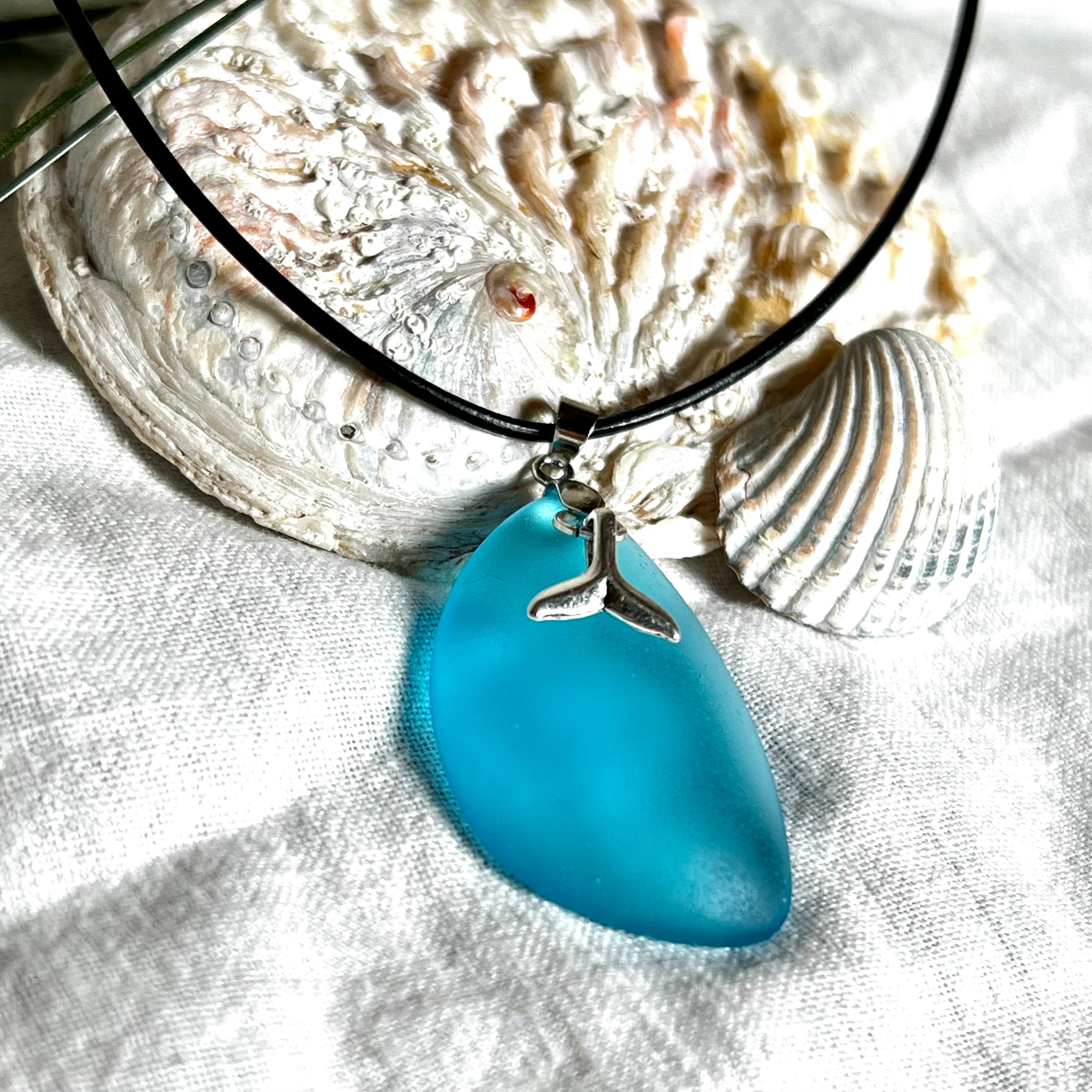 Edle Halskette „Blue Essence“ mit handgefertigtem türkisfarbenem Seeglas-Anhänger und Edelstahl-Flosse. Die Kette besteht aus schwarzer Lederkordel mit Karabinerverschluss und Verlängerung aus Edelstahl. Das matte Seeglas reflektiert Licht wie Meereswasser – ideal für Strandliebhaber und Naturliebhaber.