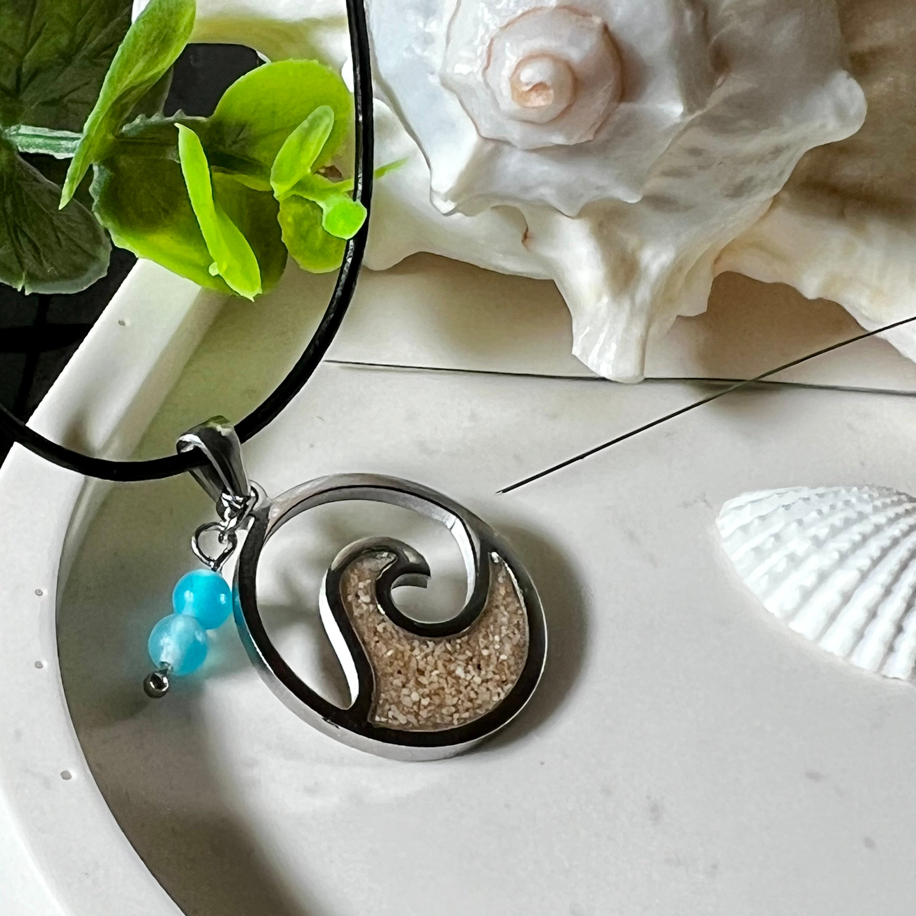 Wave Spirit – runder Edelstahlanhänger mit echter Sandfüllung in Harz eingebettet, inspiriert von einer stilisierten Welle. Verziert mit zwei blauen Achatperlen. Der Anhänger (ca. 2,5 cm Durchmesser) hängt an einer 45 cm langen Lederkette mit Edelstahlverschluss und 5 cm Verlängerungskette. Maritimer Look trifft moderne Eleganz – ein handgefertigtes Schmuckstück für Meerliebhaber