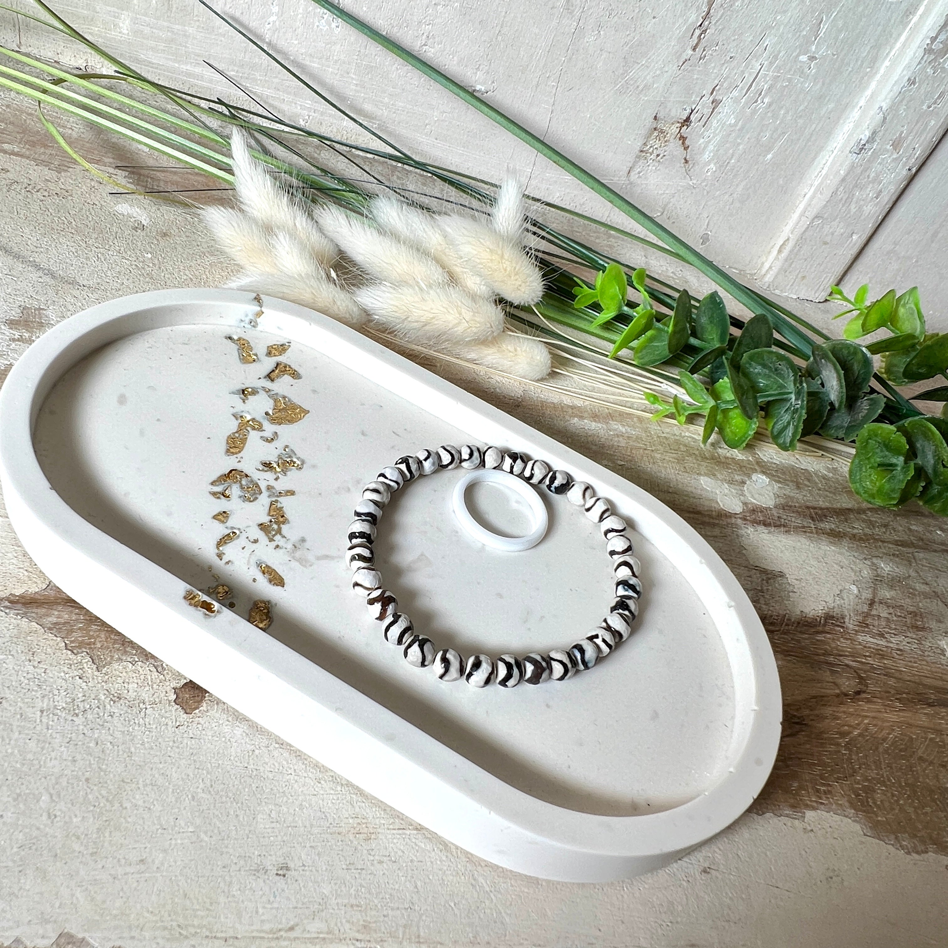 Handgefertigtes ovales Schmucktablett aus Jesmonite mit schimmernden Blattgold-Akzenten. Die Rückseite ist mit rutschfesten Korkpunkten ausgestattet. Ideal zur stilvollen Aufbewahrung von Schmuck, Schlüsseln oder kleinen Accessoires auf Kommoden und Nachttischen. Helles Design mit natürlicher Oberfläche und modernem Look. Unikat in feiner Handarbeit gefertigt – ohne Deko geliefert.