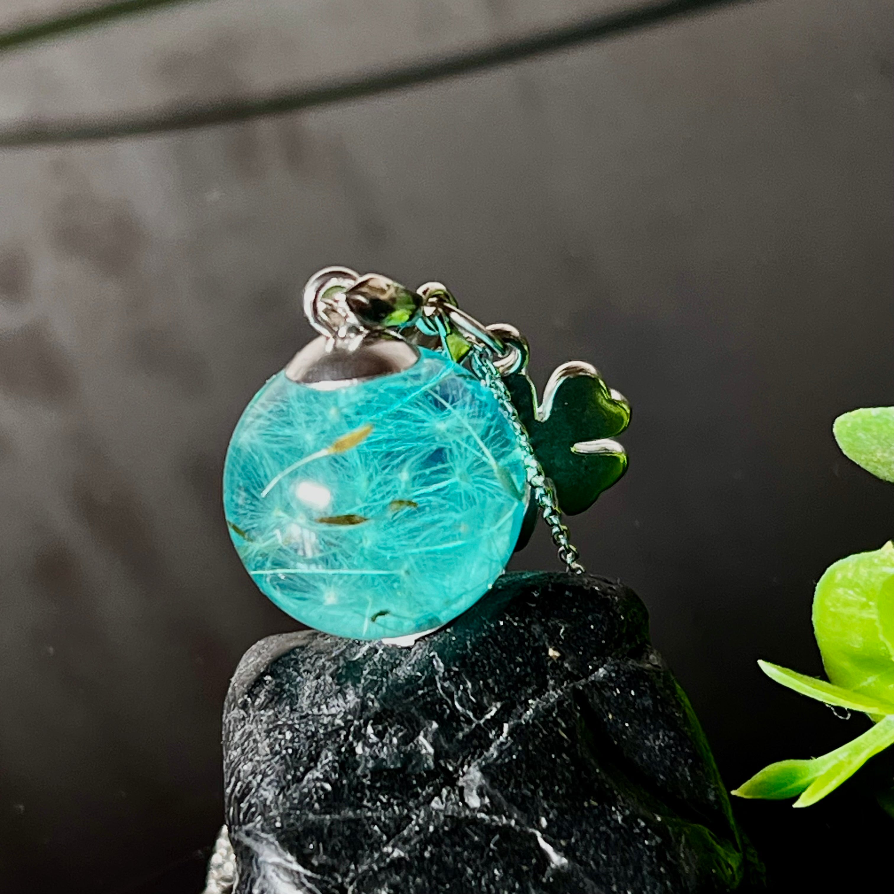Eine handgefertigten Halskette mit türkiser Harzkugel, in der echte Pusteblumensamen eingeschlossen sind. Ergänzt durch einen glänzenden Edelstahl-Kleeblatt-Charm und einer feinen rhodinierten 925 Silberkette. Der Anhänger symbolisiert Glück, Leichtigkeit und neue Wege – ein emotionales Schmuckstück mit Bedeutung.