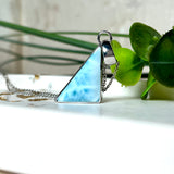 Larimar Dreieck-Anhänger in rhodiniertem 925 Sterling Silber mit filigraner Silberkette, fotografiert auf weißem Untergrund mit Goldflocken und grüner Pflanze – eleganter Edelsteinschmuck mit maritimem Flair.