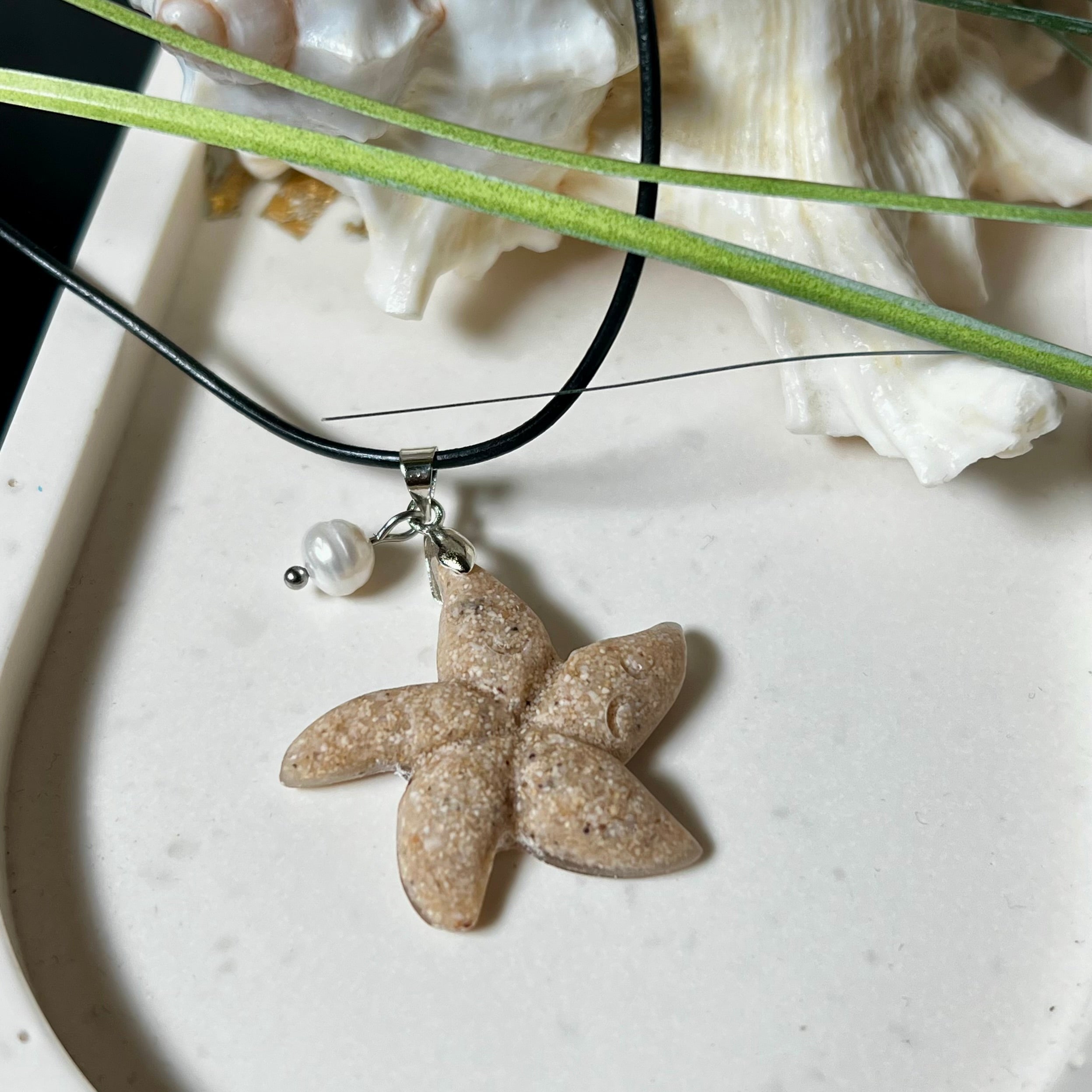 Handgefertigter Anhänger in Seesternform aus echtem Meersand und transparentem Harz. Verziert mit einer natürlichen Süßwasserperle. Edelstahlöse, Lederkette mit Karabinerverschluss (45 cm + 5 cm Verlängerung). Maritimer Schmuck im Unikatcharakter – inspiriert vom Meer und handgefertigt mit Liebe zum Detail. Perfektes Geschenk für Strandliebhaber.