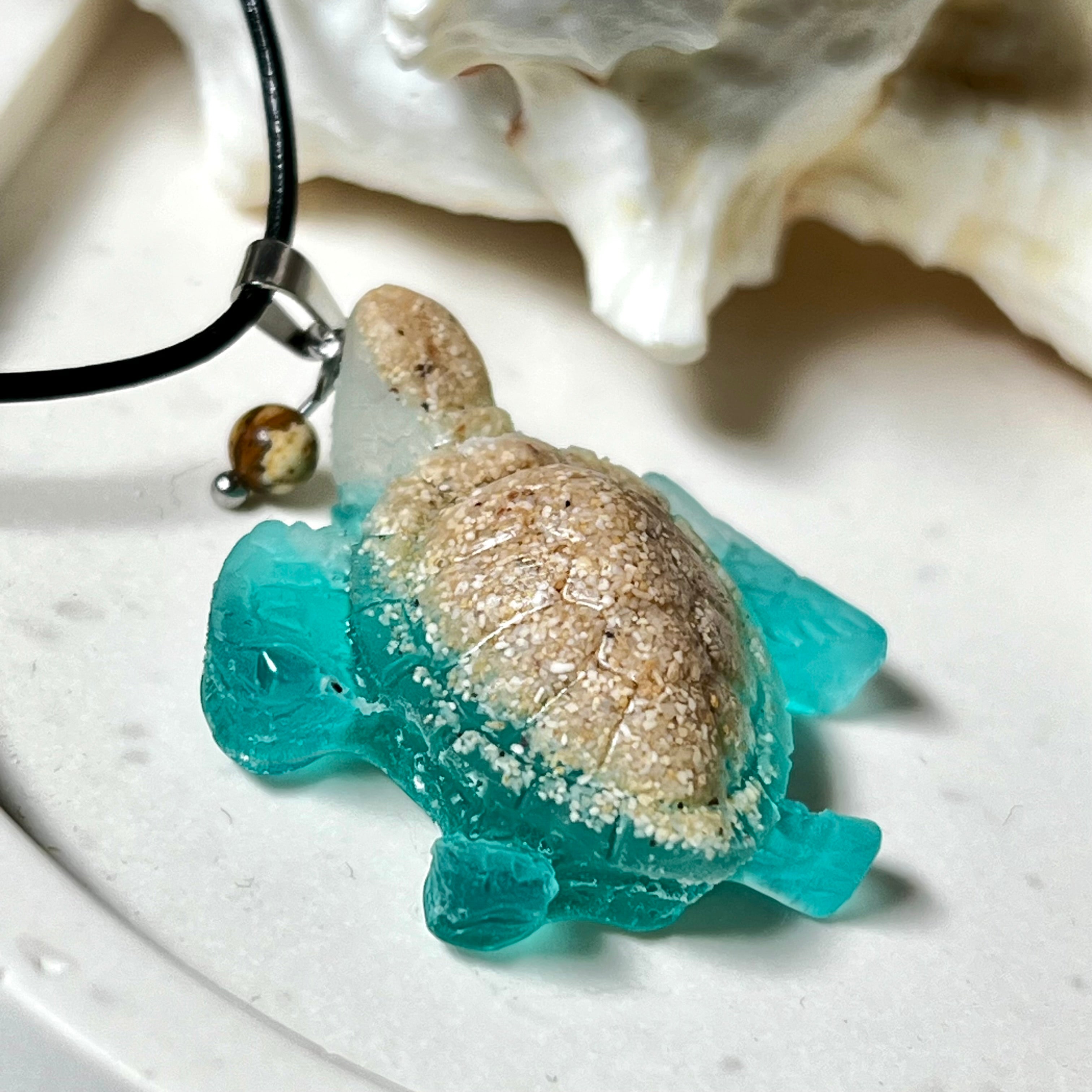 Handgefertigter Schildkröten-Anhänger Turtle Harmony aus echtem Meersand und transparent-türkisfarbenem Harz, kombiniert mit einer kleinen Jaspisperle. Symbol für Weisheit, Schutz und Harmonie. Mit Edelstahl-Anhängerschlaufe und Lederkette (45 + 5 cm). Maritimer Schmuck als Unikat, ideal als liebevolles Geschenk oder stilvolles Accessoire für den Alltag.