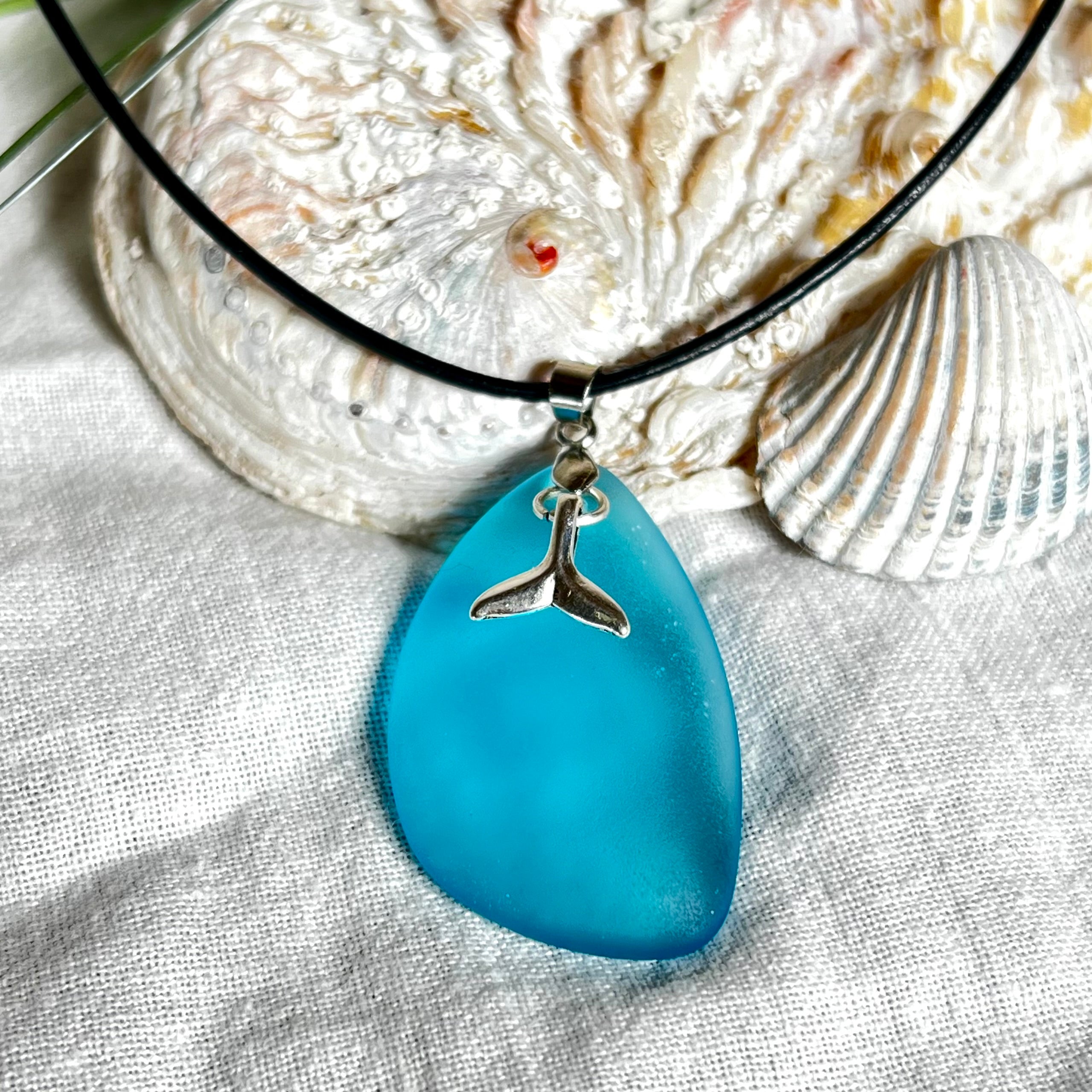Edle Halskette „Blue Essence“ mit handgefertigtem türkisfarbenem Seeglas-Anhänger und Edelstahl-Flosse. Die Kette besteht aus schwarzer Lederkordel mit Karabinerverschluss und Verlängerung aus Edelstahl. Das matte Seeglas reflektiert Licht wie Meereswasser – ideal für Strandliebhaber und Naturliebhaber.
