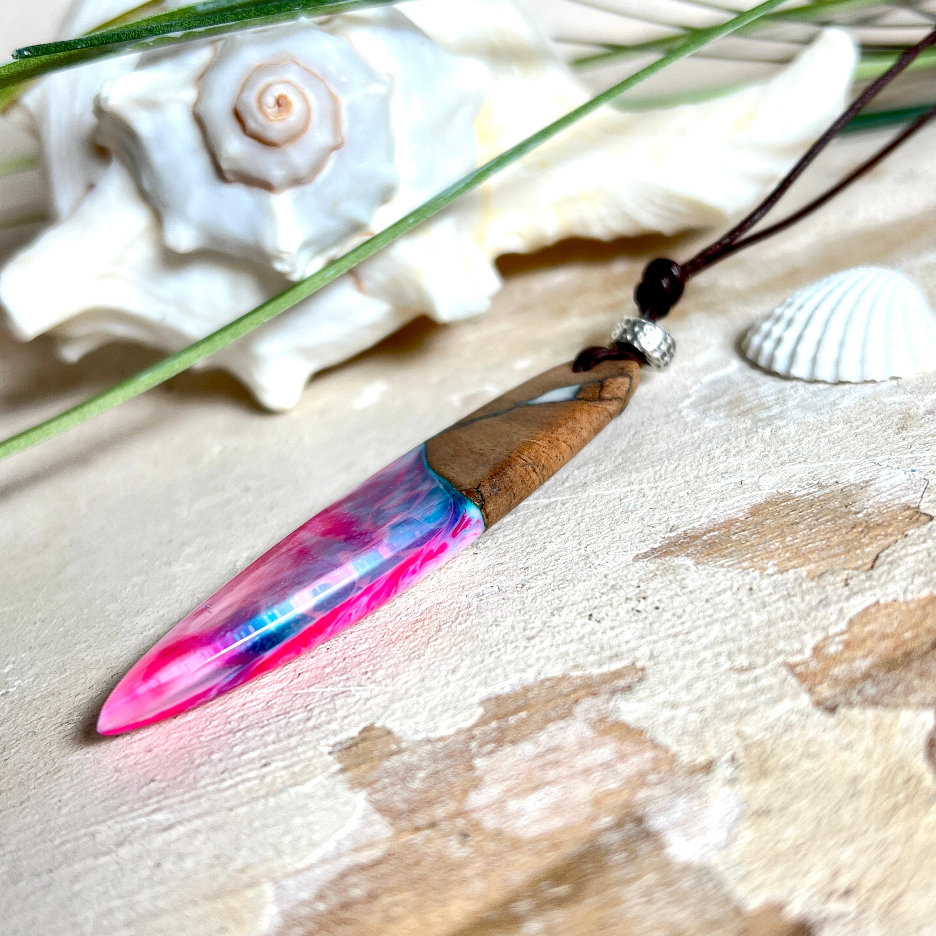 Handgefertigter Surfbrett-Anhänger aus Nussbaumholz und fluoreszierendem Epoxidharz in Pink, Blau und Weiß. Der Anhänger leuchtet zart bei Schwarzlicht und wird an einem verstellbaren Lederband (25–44 cm) mit Holzperlen getragen. Ein maritimes Unikat – ideal für Surfer und Naturliebhaber.