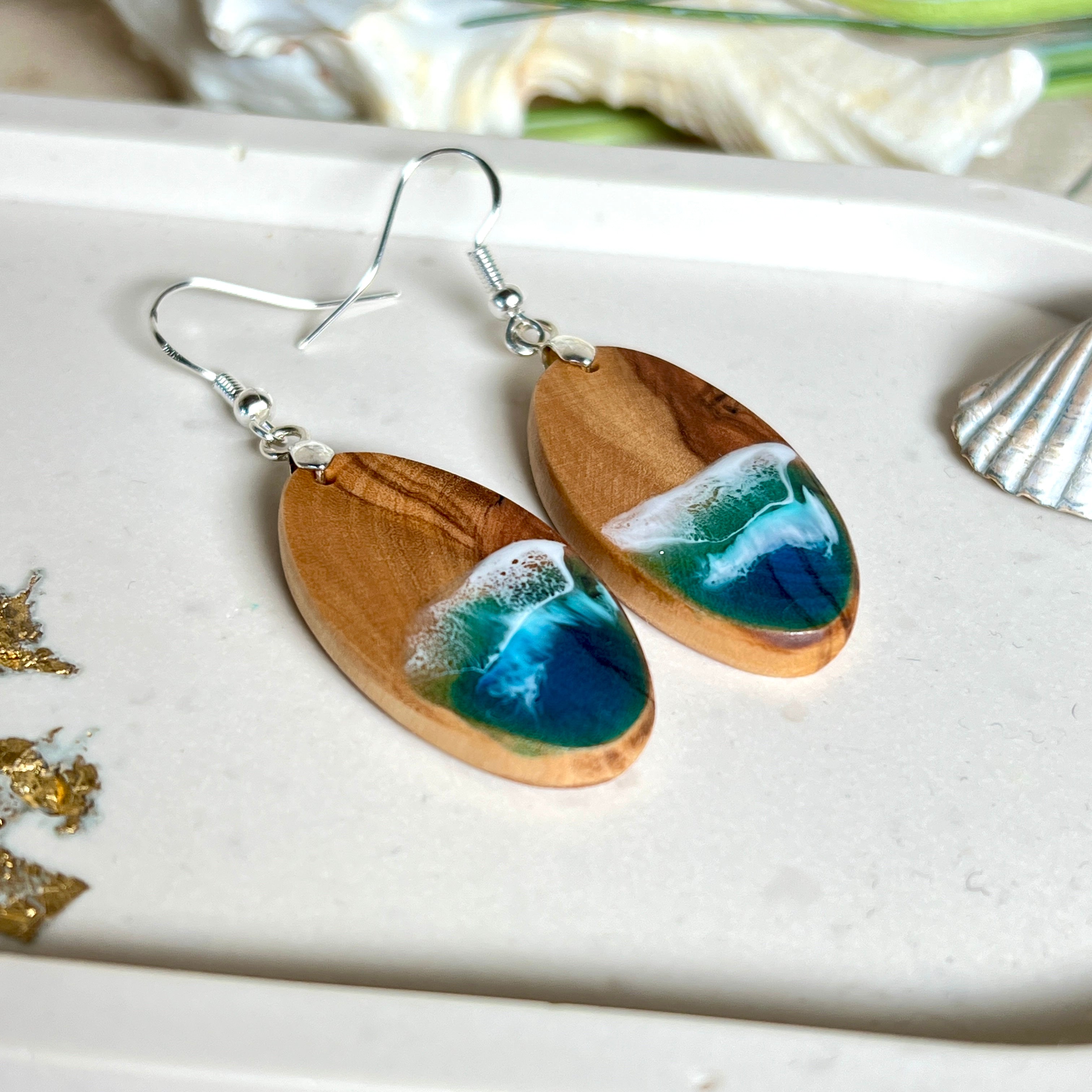 Handgefertigte Ohrringe „Wave Harmony“ aus Olivenholz und Epoxidharz in Blau-, Türkis- und Weißtönen. Das Design erinnert an Meereswellen am Strand. Jeder Ohrring ist ein Unikat mit individueller Maserung. Versilberte, nickelfreie und bleifreie Anhängerschlaufe, 925 Silber Ohrhaken. Maritimer Schmuck – perfekt für Meerliebhaber und als edles Geschenk.