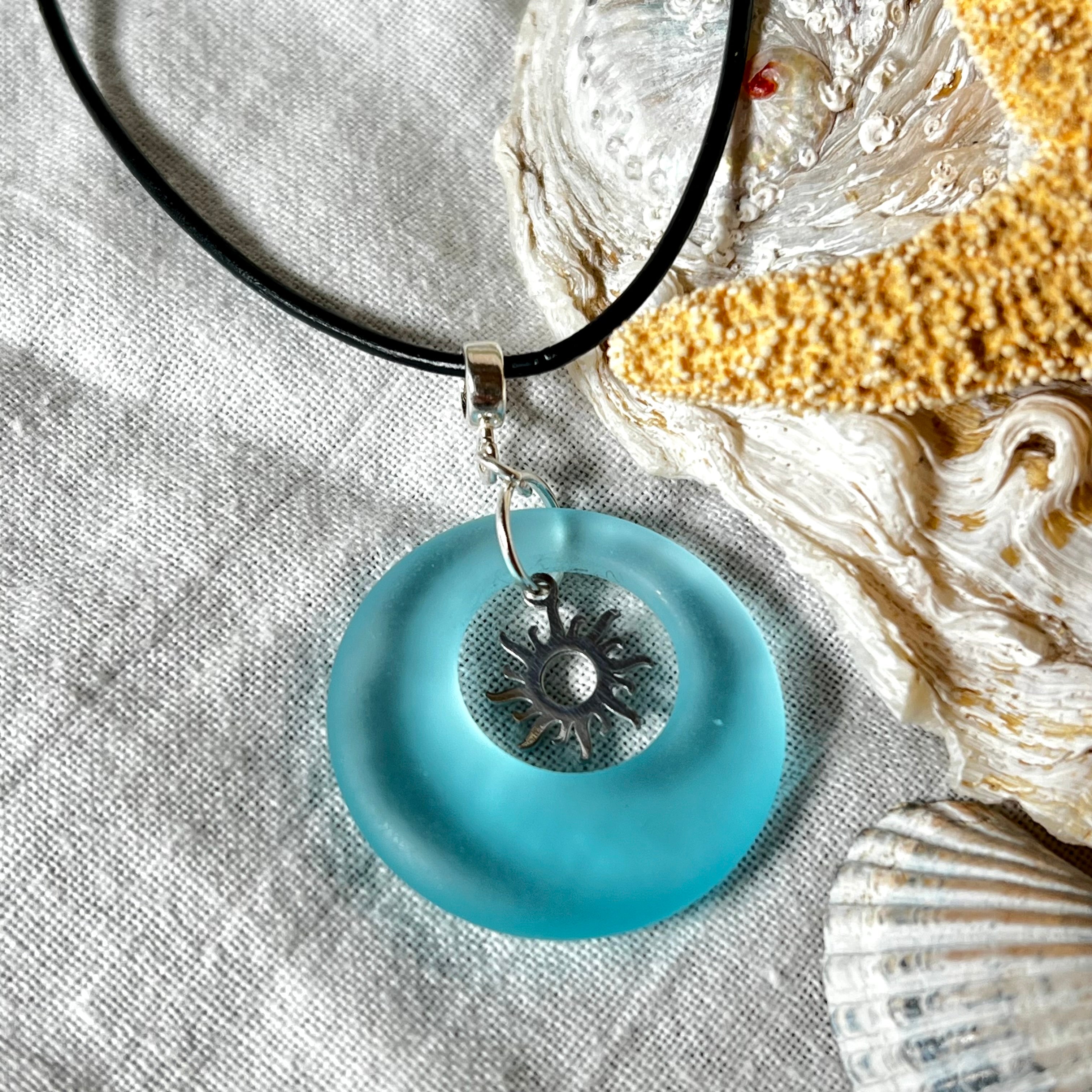 Handgefertigte Halskette mit Donut-Anhänger aus türkisfarbenem Seeglas, kombiniert mit einem filigranen Sonnen-Charm aus Edelstahl. Die Kette besteht aus einer schwarzen Lederschnur (1,5 mm) mit Karabiner und Verlängerungskette aus Edelstahl. Maritimes Schmuckstück für Meerliebhaberinnen und Sommerfreunde. Unikat mit stilvoller Ausstrahlung.