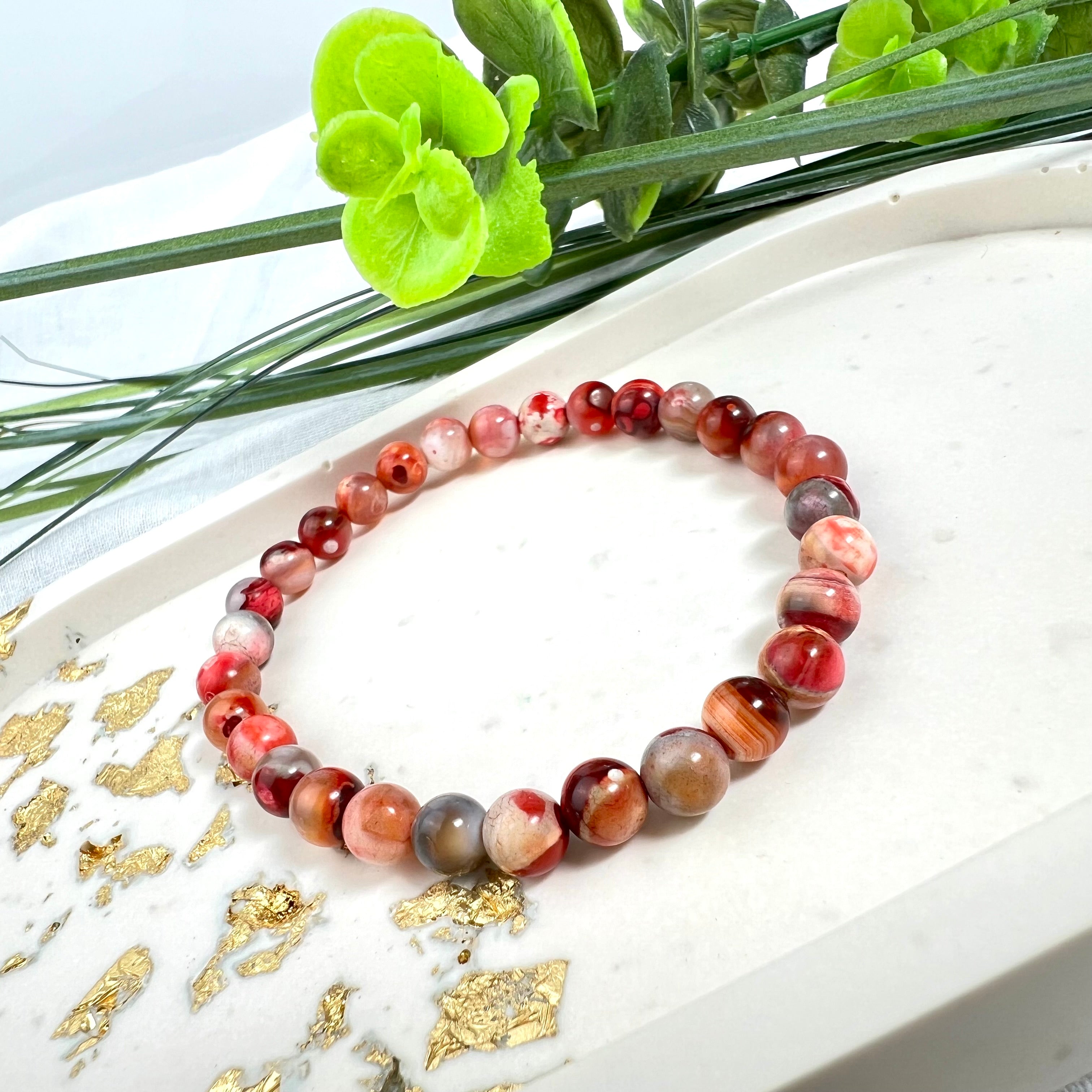 “Handgefertigtes Rot-Pink-Achat-Armband auf hellem Untergrund mit goldenen Akzenten und grünen Blättern im Hintergrund – ideal für Schmuckliebhaber, Energiearbeit und als Geschenk.”