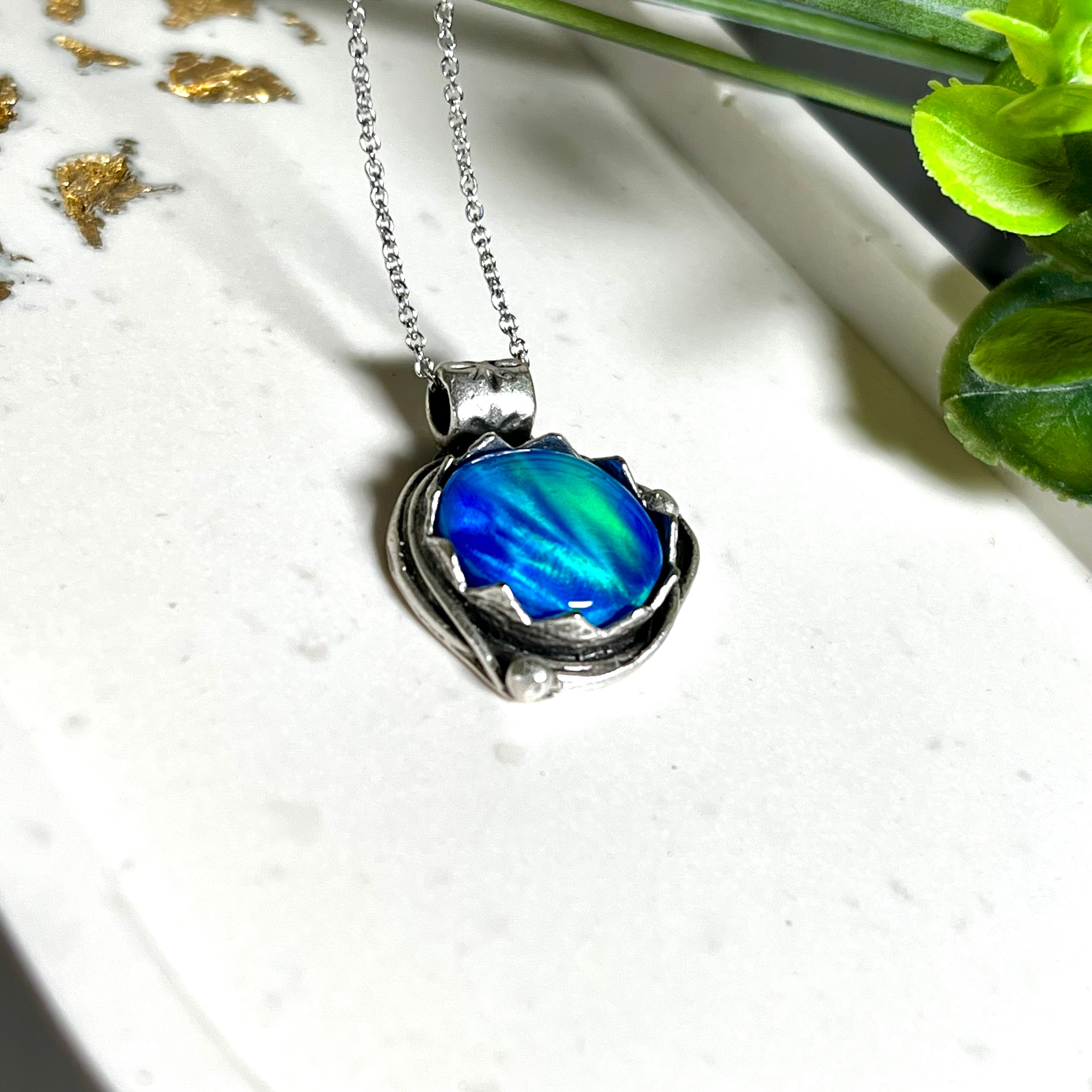 Handgemachter Anhänger mit rundem Cabochon aus Epoxidharz und schimmerndem synthetischem Opal. Die Farben changieren je nach Lichteinfall in Blau, Türkis und Grün. Der Opal sitzt in einer antik versilberten Fassung aus Messing (blei- & nickelfrei). Geliefert mit einer rhodinierten 925 Silberkette (45 cm). Jedes Stück ein Einzelstück – perfekt als stilvolles Geschenk oder persönlicher Glücksbringer.